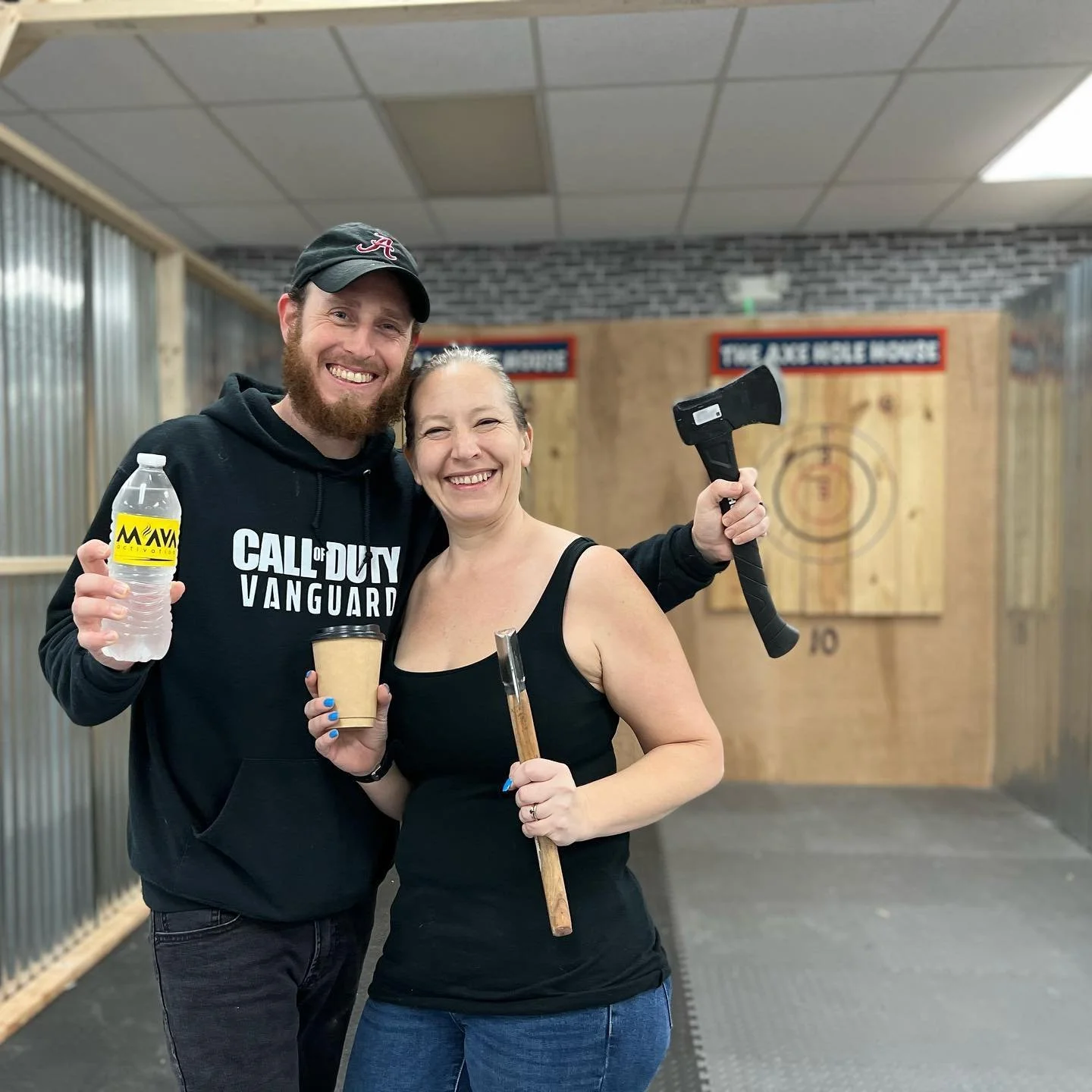 A man and woman smiling at a shooting range. The man is holding a water bottle and a cup, the woman is holding a stick and an axe, and she has an axe in her hand. They are standing in front of an archery target on a wooden wall.
