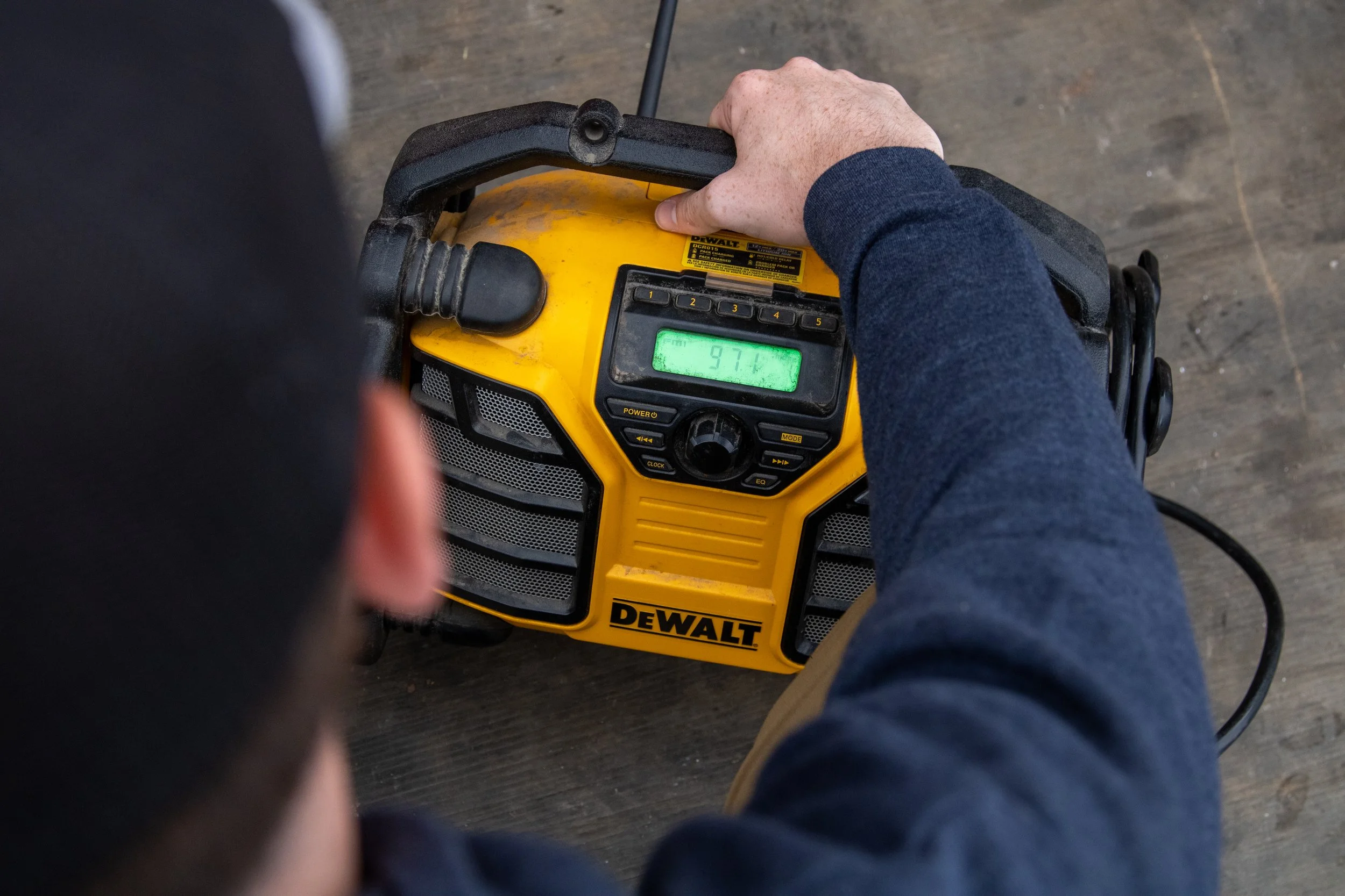 A person's hand resting on a yellow DeWalt power tool with a digital screen displaying 97.1. The person is wearing a blue long sleeve shirt and is seated on a dark wooden surface.