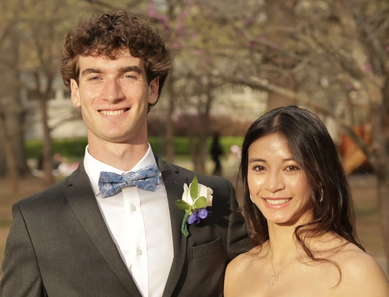 A young man in a tuxedo with a blue bow tie and a woman in a strapless dress, both smiling outdoors during daytime with trees in the background.