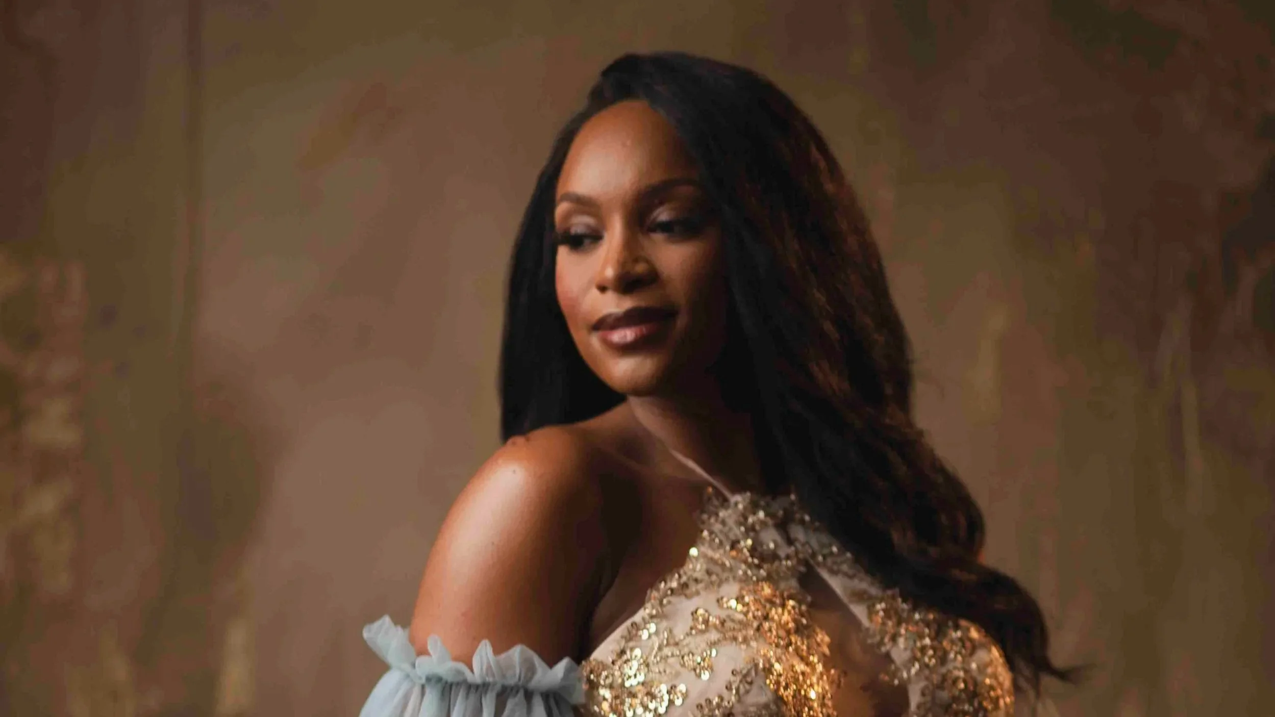 A woman with long black hair wearing an elegant dress with gold sequins, posing indoors with softly lit background.