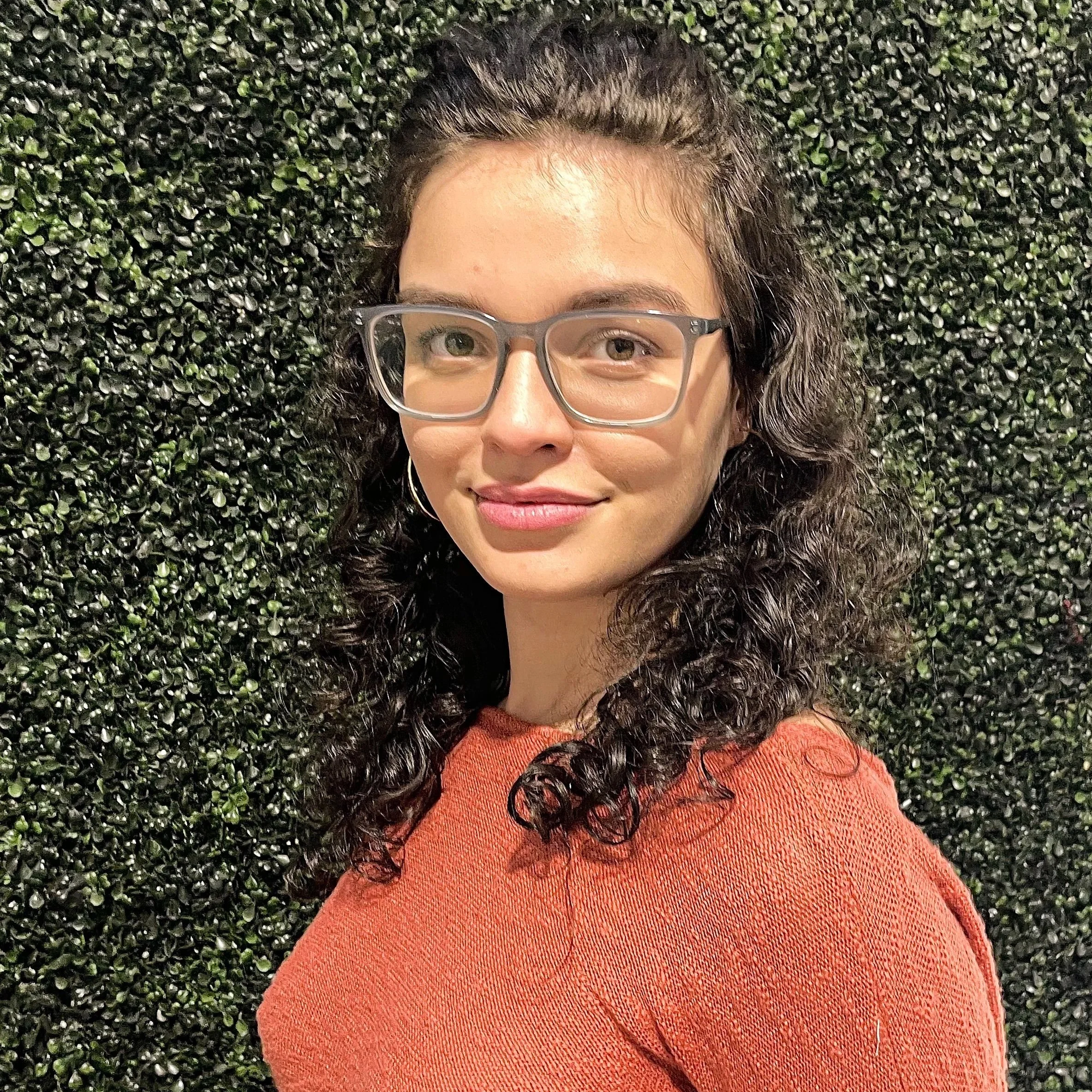 A woman with curly dark hair wearing glasses and an orange sweater stands in front of a green leafy wall.