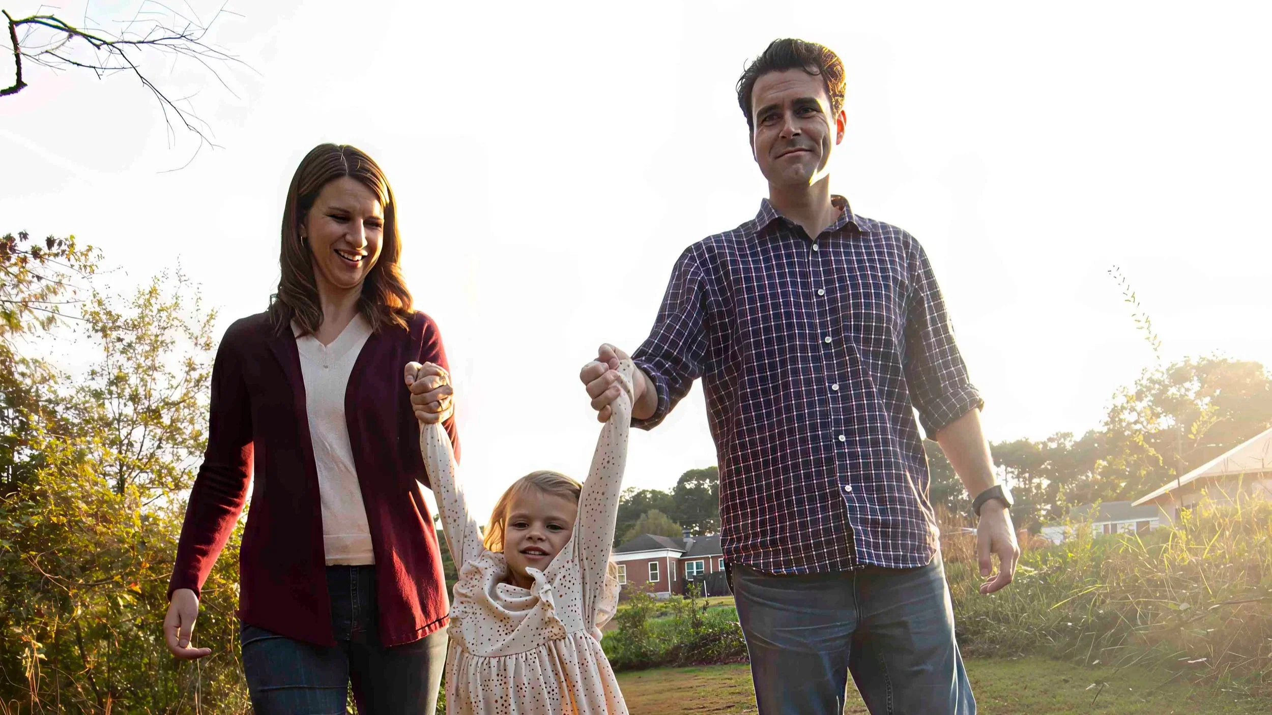 A family of four, including a woman, a man, and two children, are smiling and holding hands outdoors on a sunny day, with trees and houses in the background.