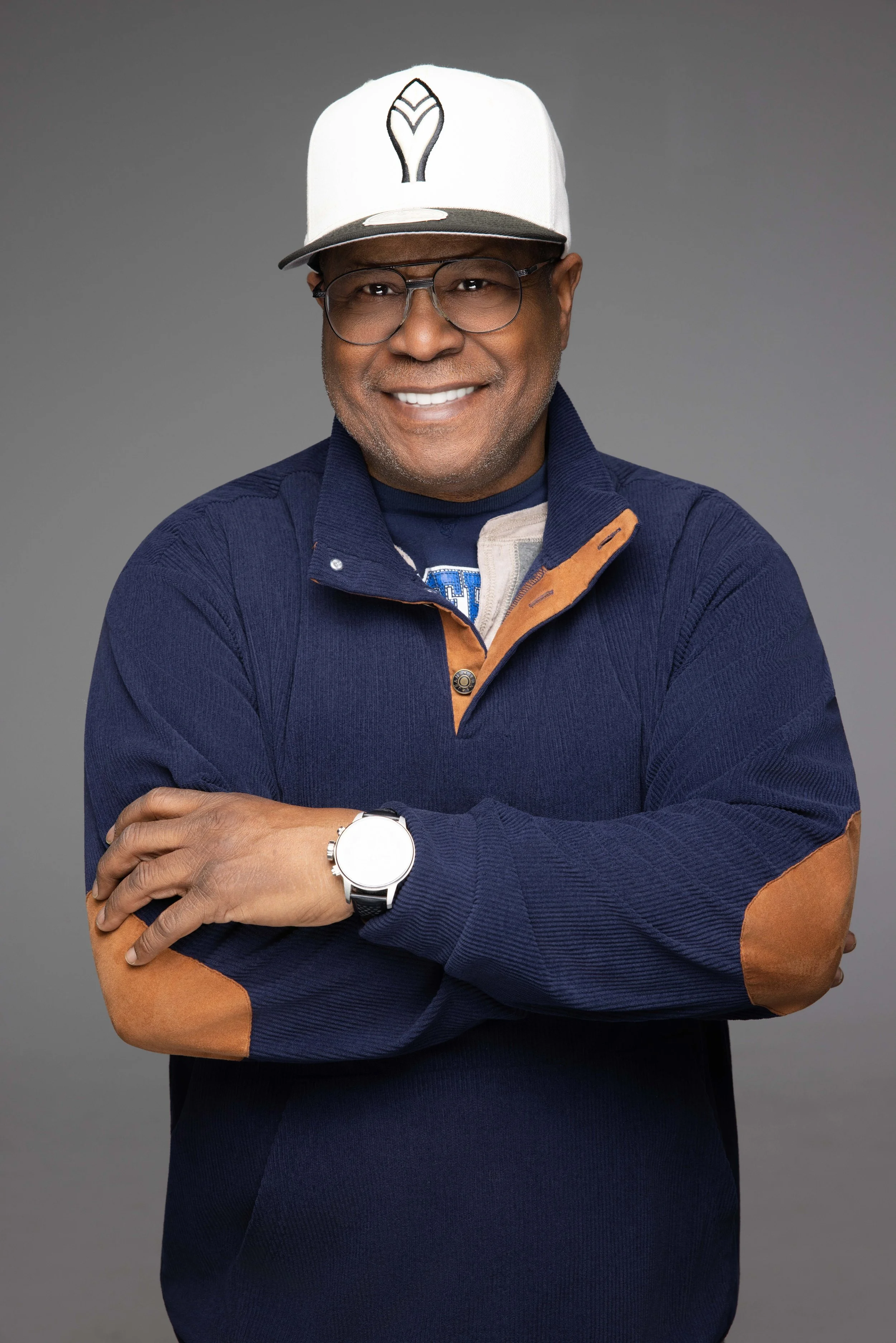 A smiling man with glasses wearing a baseball cap, a navy jacket with tan patches, and a watch, standing with crossed arms against a gray background.