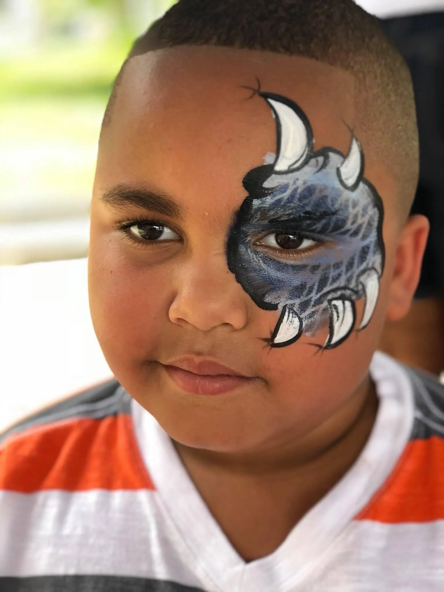 Close-up of a young boy with face paint depicting a roaring black and gray animal with white teeth, likely a bear or tiger, covering part of his face, with short hair and wearing a striped shirt.