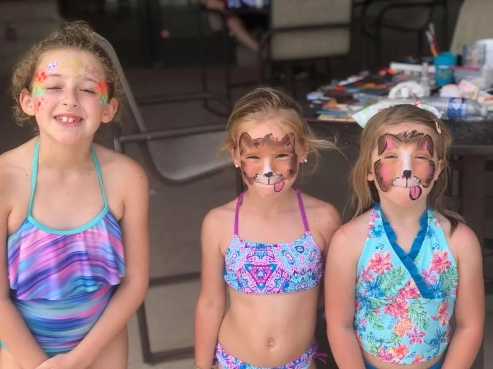 Three young girls with face paint, standing together outdoors in swimsuits, smiling and enjoying a sunny day.