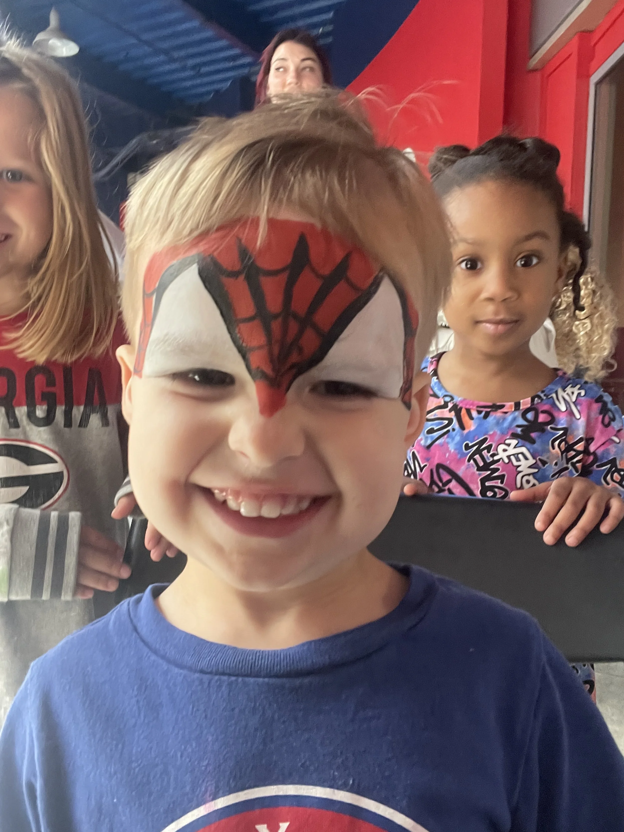 A young boy with a face painted as Spiderman smiling for the camera, with three other children and a woman in the background.