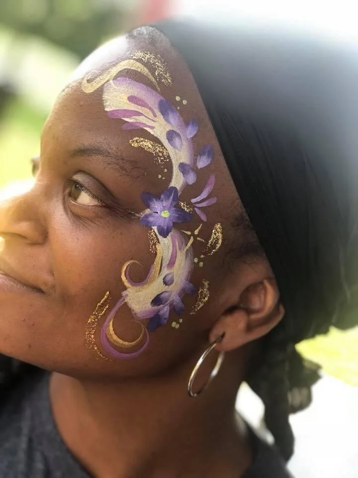 A woman with face paint featuring purple flowers and gold accents on her left cheek, wearing a black headscarf and hoop earrings.
