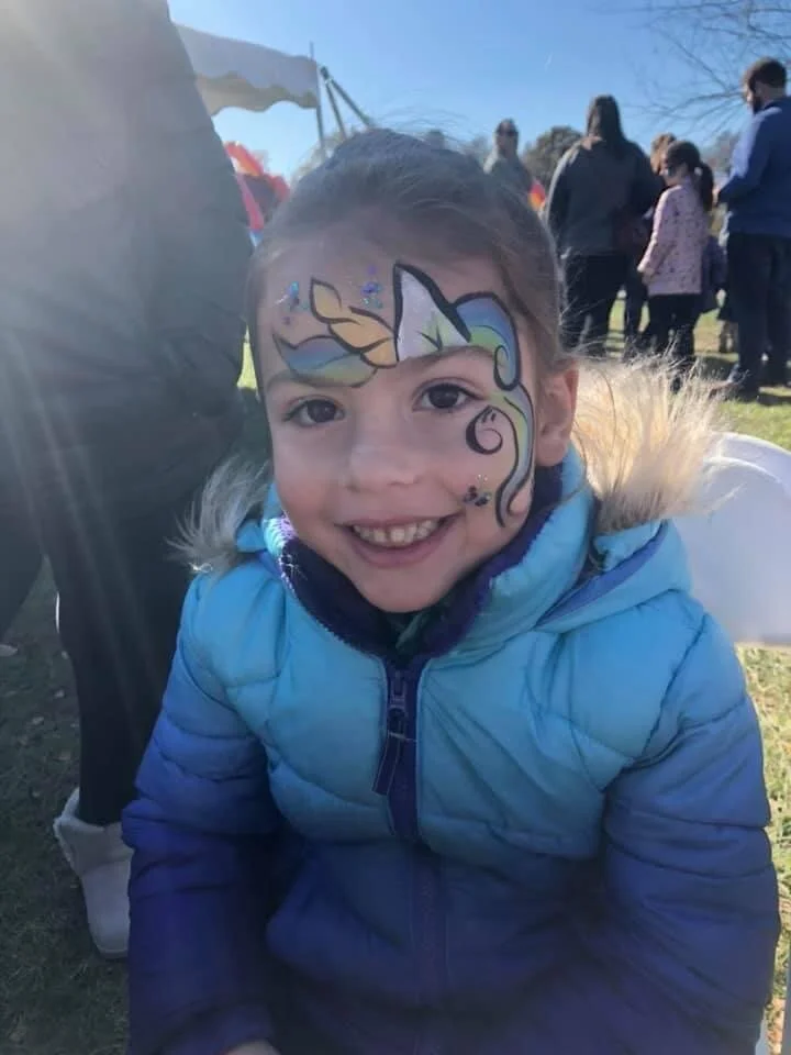 A smiling young girl with face paint of a colorful unicorn on her face, wearing a blue jacket, at an outdoor event on a sunny day.