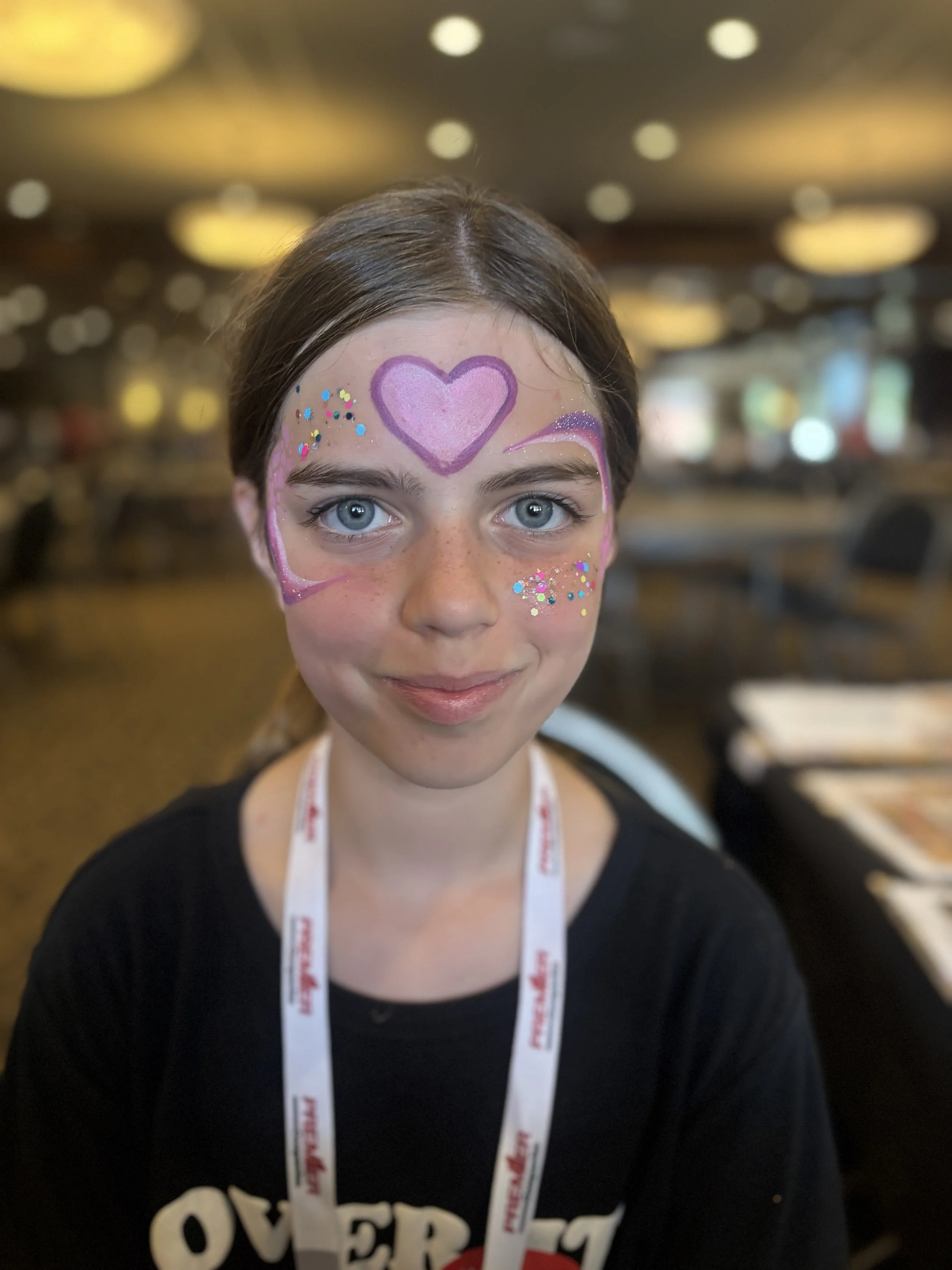 Girl with face paint featuring a purple heart on her forehead, pink and purple lines around her eyes, and colorful glitter on her cheeks, in a room with warm lighting.