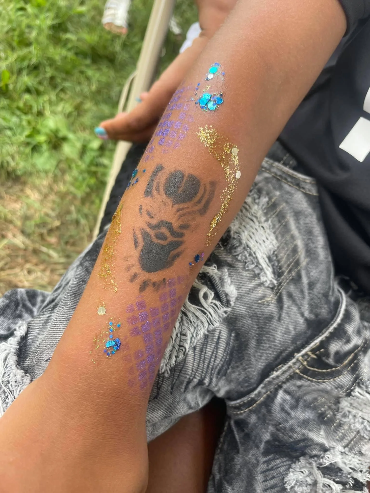 Temporary tattoo of a tiger's face with purple and blue glitter embellishments on an arm with gold glitter accents, next to ripped gray denim shorts.