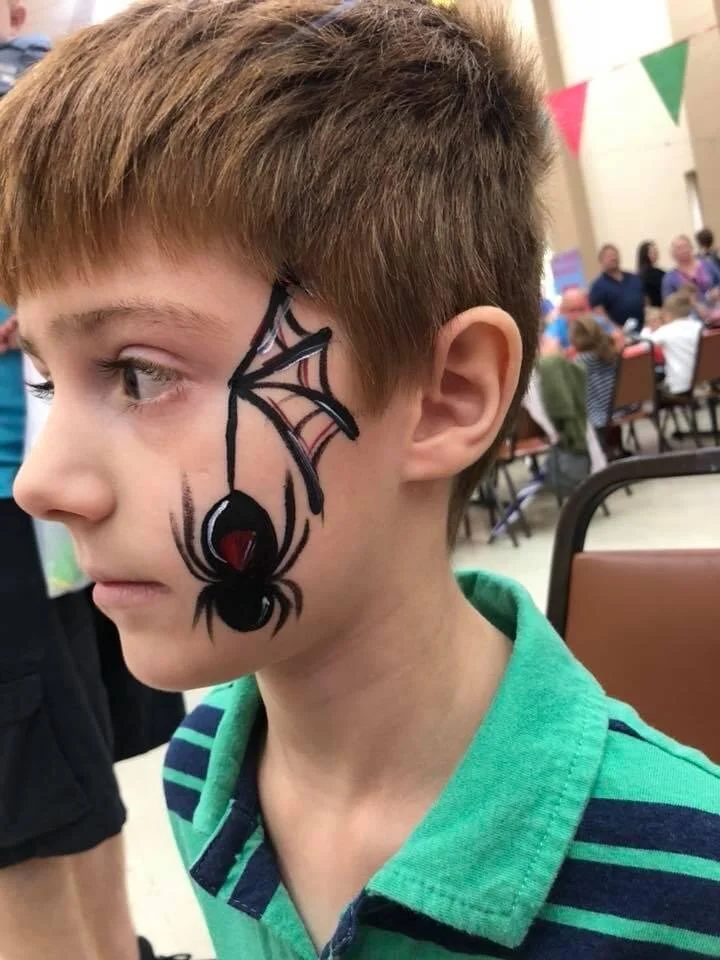 A young boy with face paint of a black spider with red eyes on his left cheek, in a crowded indoor setting with festive decorations.