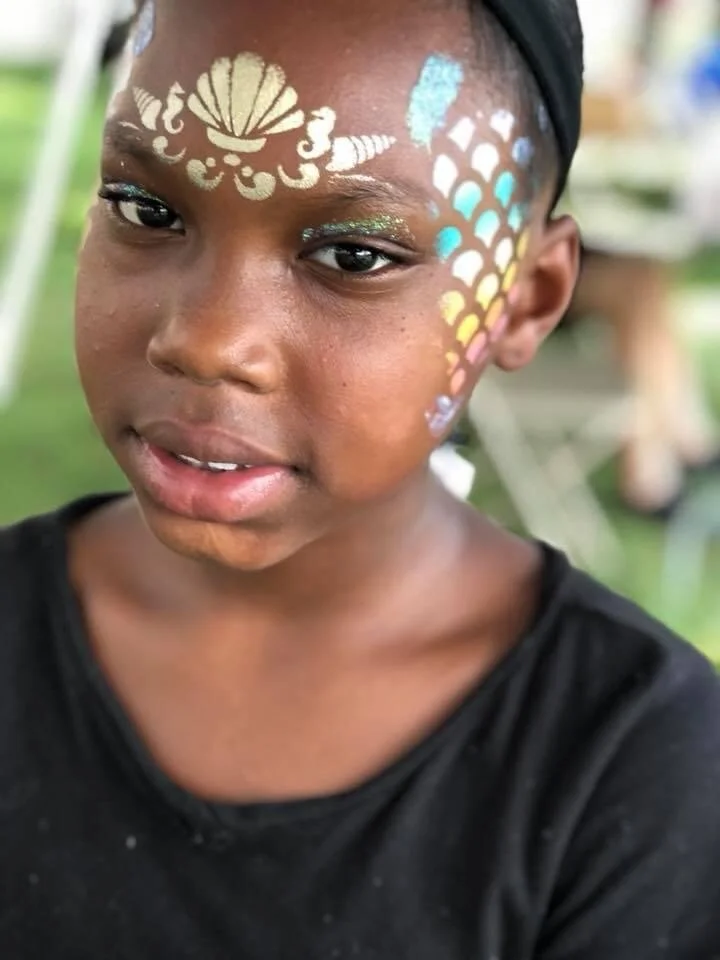 Close-up of a child with colorful face paint featuring a seashell and fish scale patterns.