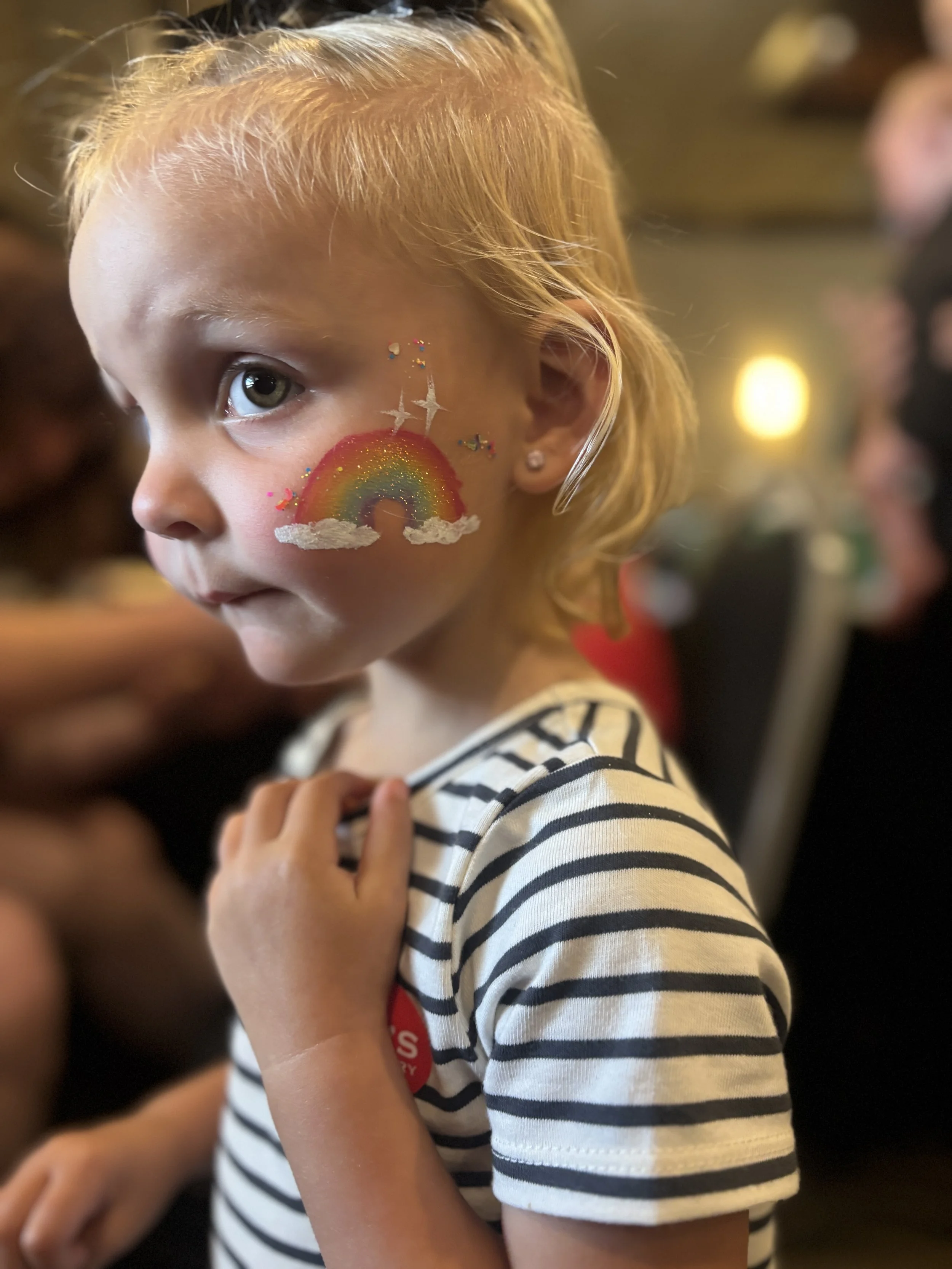 A young girl with blond hair and a striped shirt has a rainbow and clouds painted on her face.