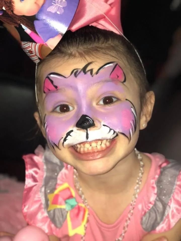 Child with face paint resembling a pink and white cat, wearing a pink princess dress and a pink party hat, smiling.