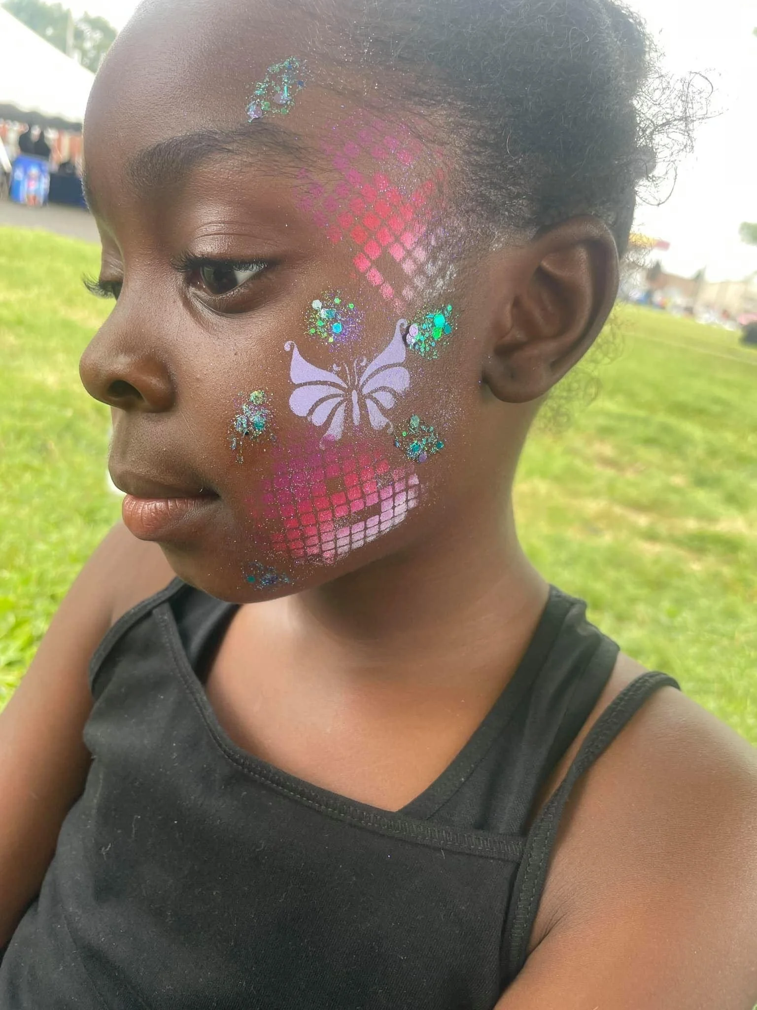 Close-up of a young girl with colorful face paint featuring a butterfly, glitter, and a pink grid pattern, outdoors on a grassy field.