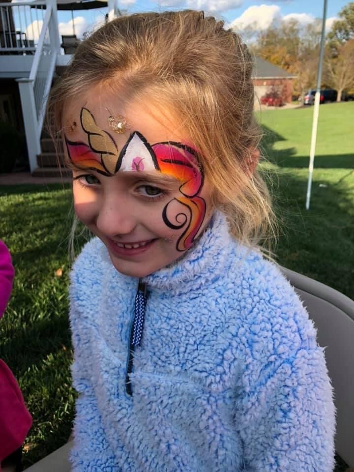 Young girl with a painted unicorn face on her face, smiling outdoors on a sunny day.