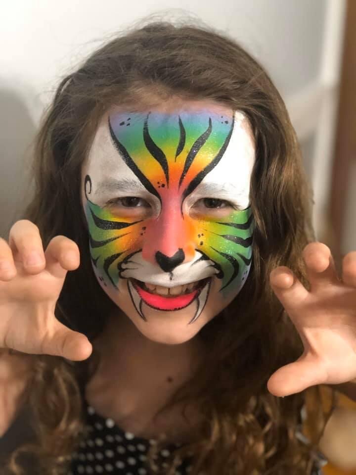 A person with face paint resembling a colorful tiger, showing claws and smiling at the camera.