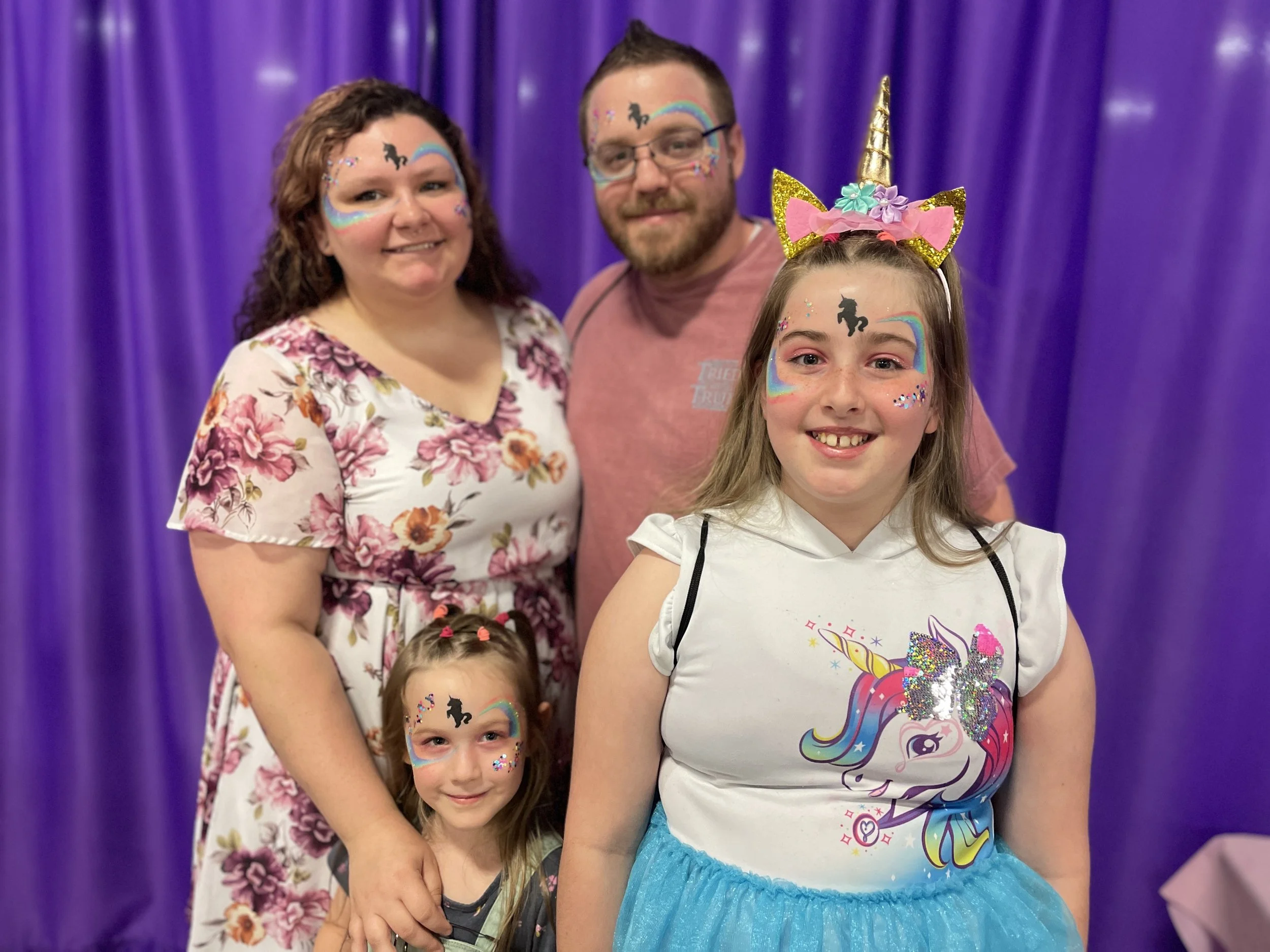 Family of four at a party with a purple curtain background. The family includes two young girls and their parents, all wearing colorful unicorn-themed face paint, accessories, and festive clothing.