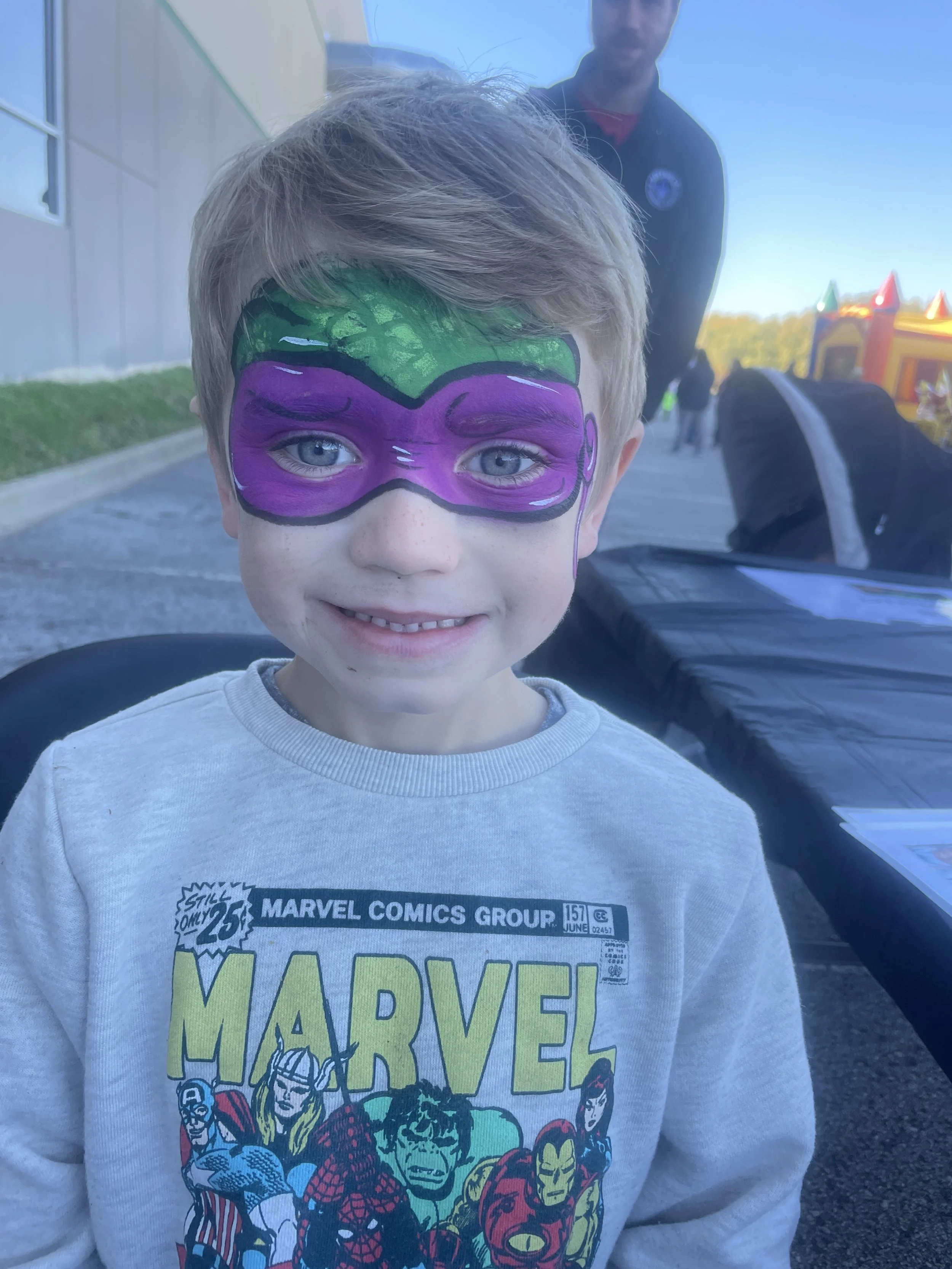 A young boy with face paint resembling a purple and green superhero mask smiling at the camera. He's wearing a Marvel Comics sweatshirt. An inflatable bounce house can be seen in the background and a man standing behind him.