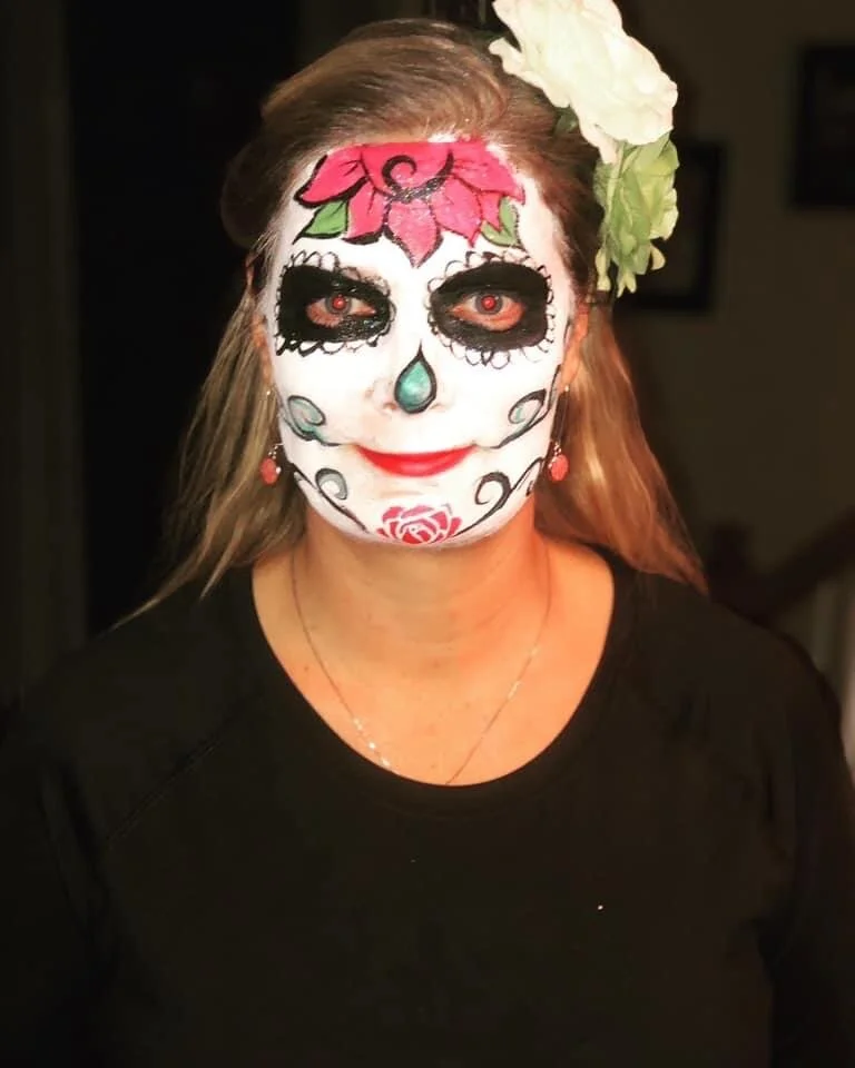 Woman with sugar skull face paint, black eye makeup, red contact lenses, green and pink floral headpiece, wearing a black shirt, and red earrings.