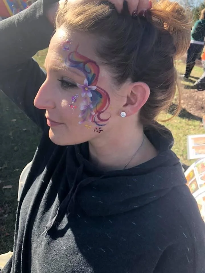 A young woman with colorful unicorn and floral face paint, wearing earrings and a black hoodie, outdoors on a sunny day.