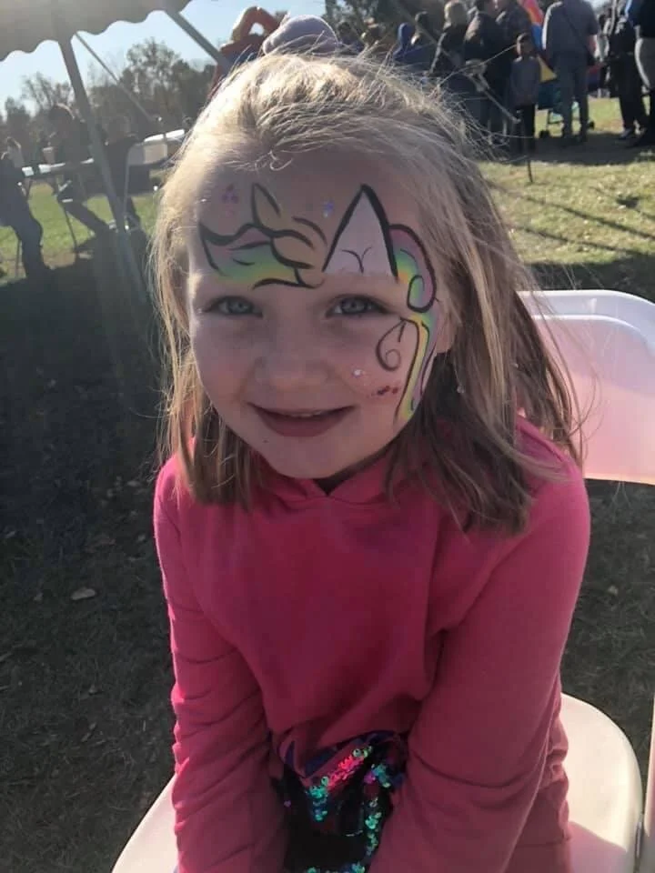 A young girl with face paint of a unicorn, smiling at an outdoor event.