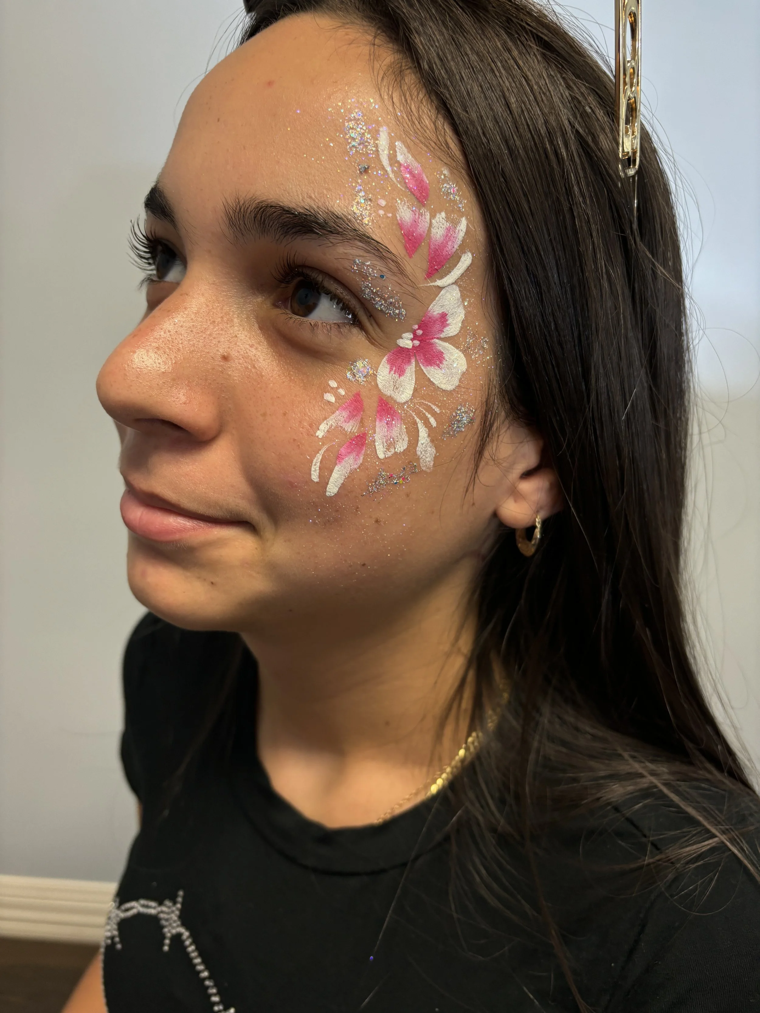 Young woman with face painted in white and pink butterfly and flower design, adorned with glitter, and wearing a black shirt, earrings, and a headband.