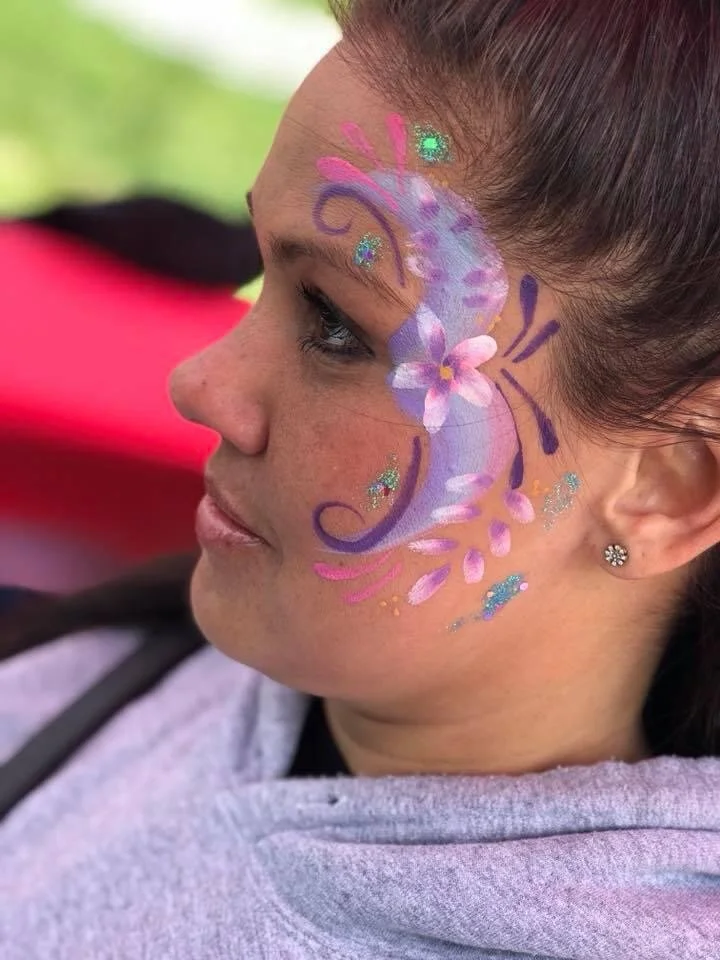 Side profile of a woman with a butterfly face painting on her cheek, featuring purple, pink, and blue colors with glitter accents.