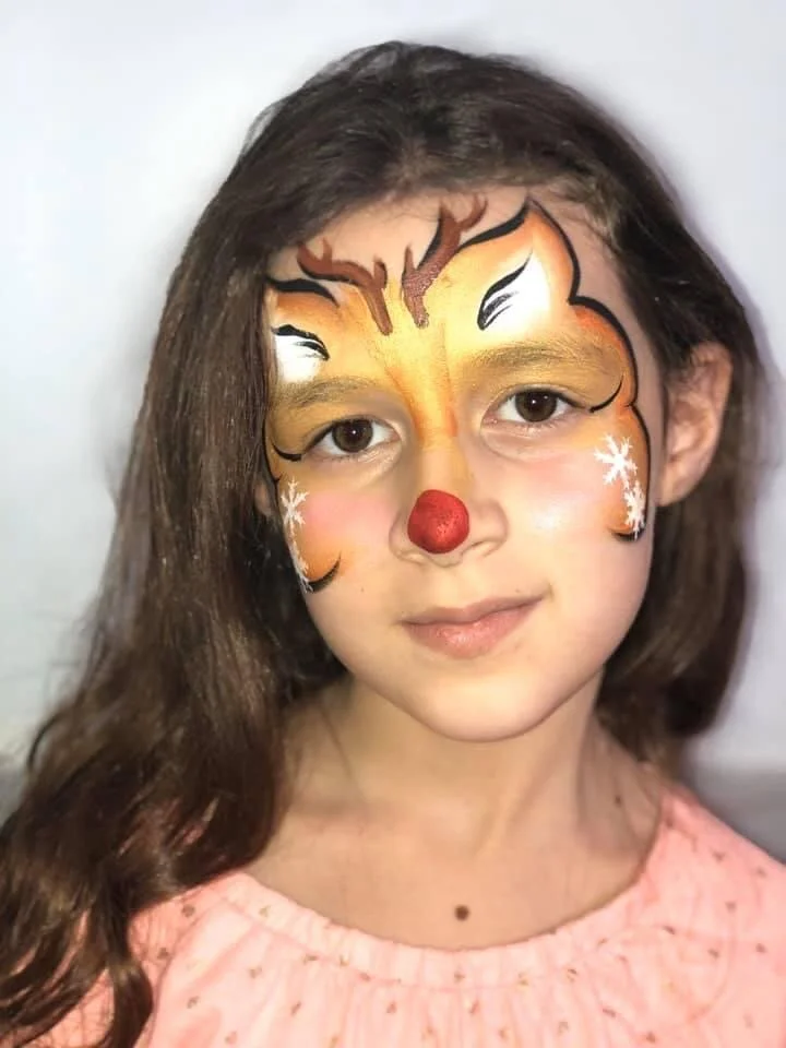 Young girl with face paint of a reindeer's face, complete with a red nose, white snowflakes, and yellow and black accents, wearing a pink top.