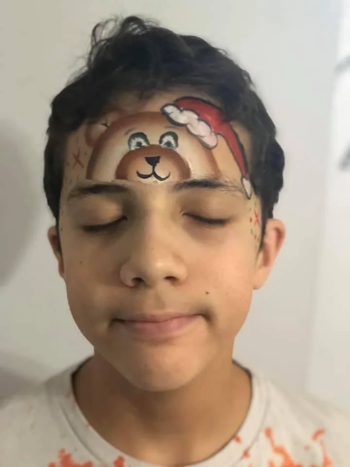A young boy with closed eyes has a Christmas-themed bear face painted on his forehead, featuring a bear wearing a Santa hat.