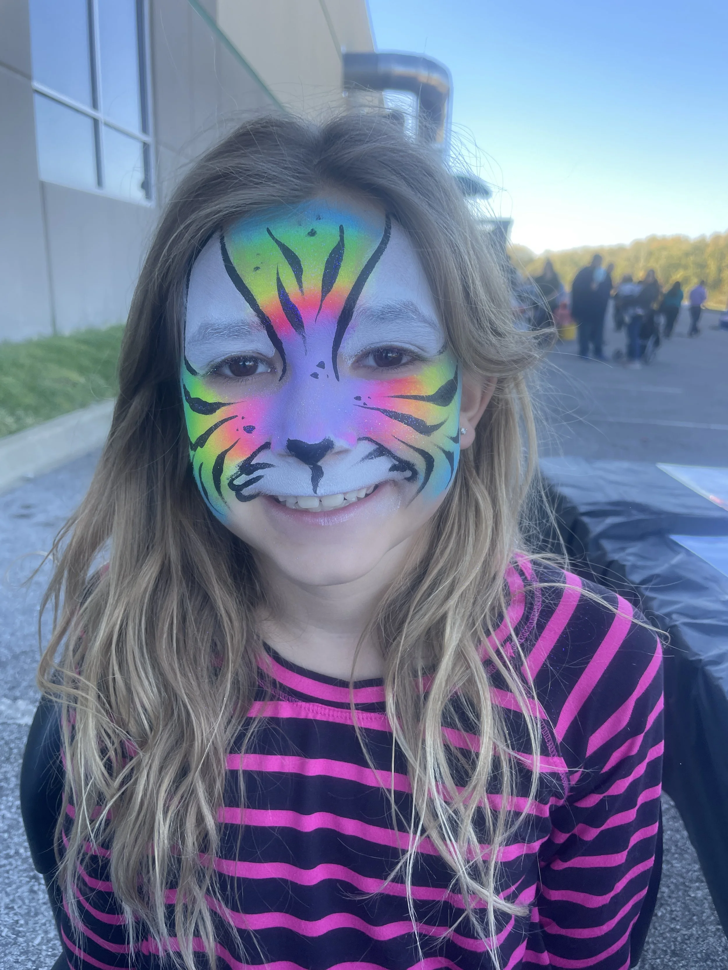 A young girl with long blonde hair and earrings, smiling while wearing colorful tiger face paint in rainbow hues on her face, outdoors near a building with other people in the background.