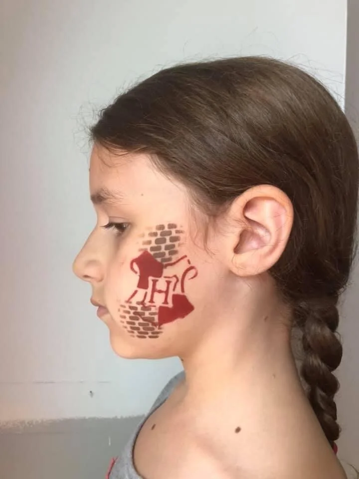 Side profile of a girl with brown hair in a braid, showing a painted design on her face of a brown mouse with a red swoosh and the letter 'H', with a brick wall pattern behind the mouse.
