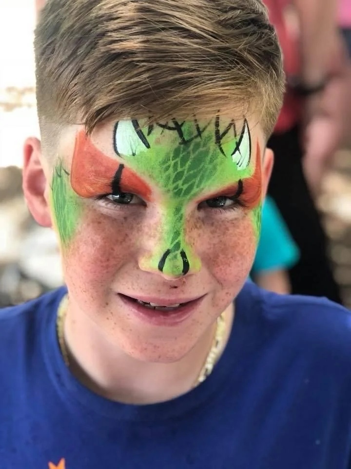 Young boy with face paint designed to look like a colorful, animated animal face, possibly a dragon or dinosaur, with green, orange, and black details.