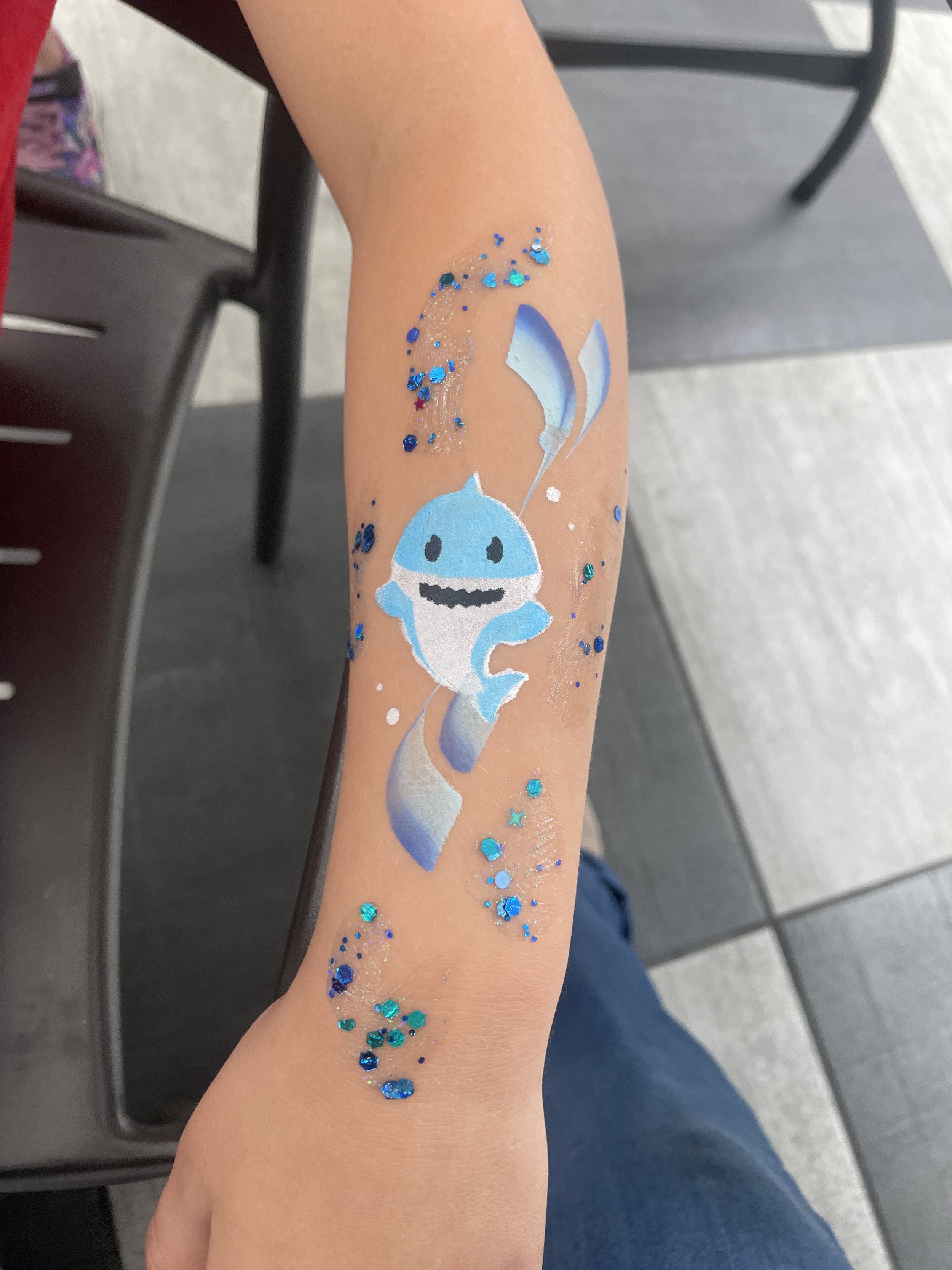Temporary tattoo of a cute, cartoon-like whale with a happy expression and a spout of water, surrounded by blue glitter, on a person's forearm.