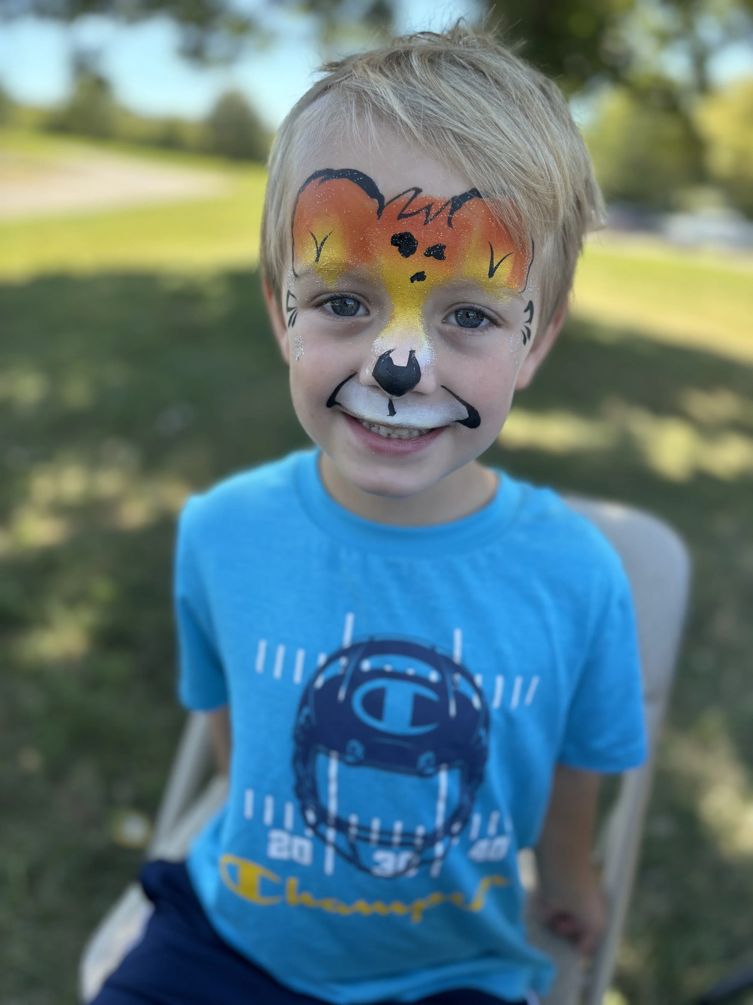 Young boy smiling with a painted fox face on his face, outdoors on a sunny day.