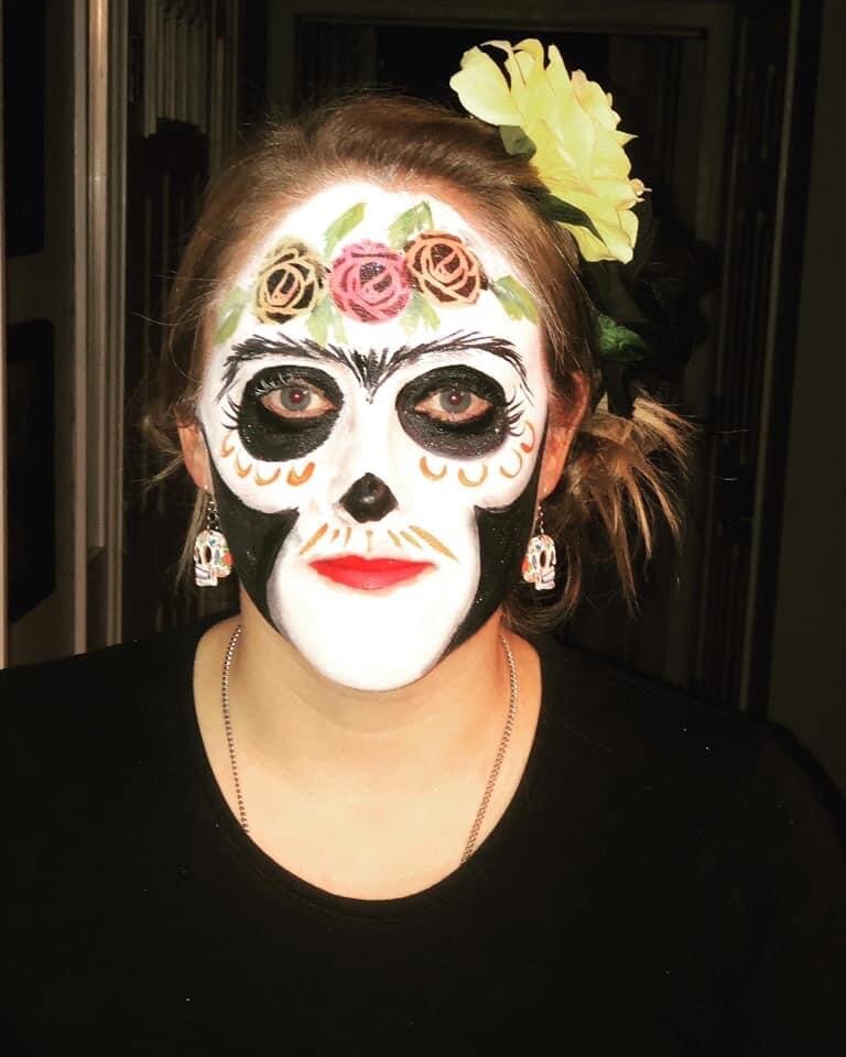 A woman with 'calavera' (sugar skull) face paint, featuring intricate floral and skull motifs, black eye makeup, red lipstick, earrings, a flower in her hair, wearing a black top.