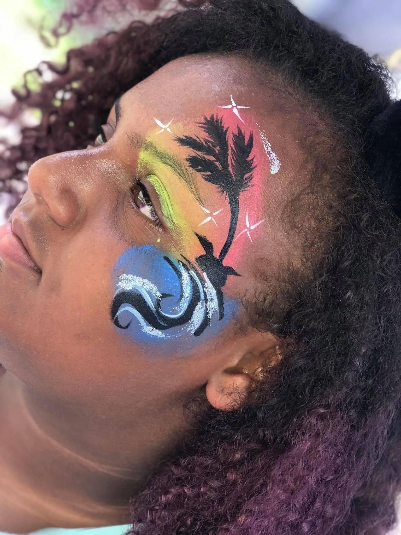 Close-up of a woman with colorful face paint featuring a black silhouette of a plant, blue waves, and sparkles around her eye and forehead.