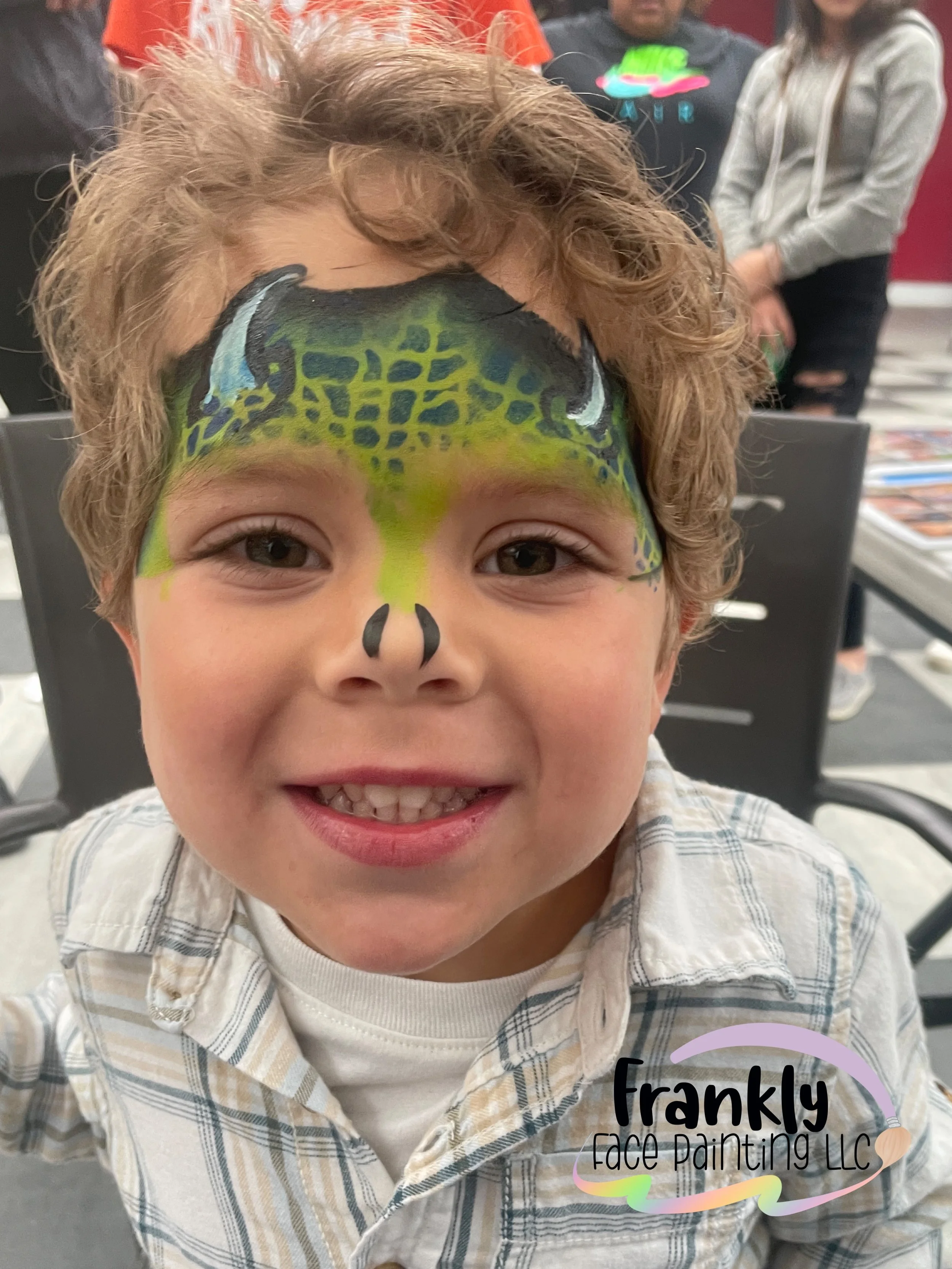 A young boy smiling with face paint that resembles a green and blue dragon, with detailed scales and eye features, at a face painting event.