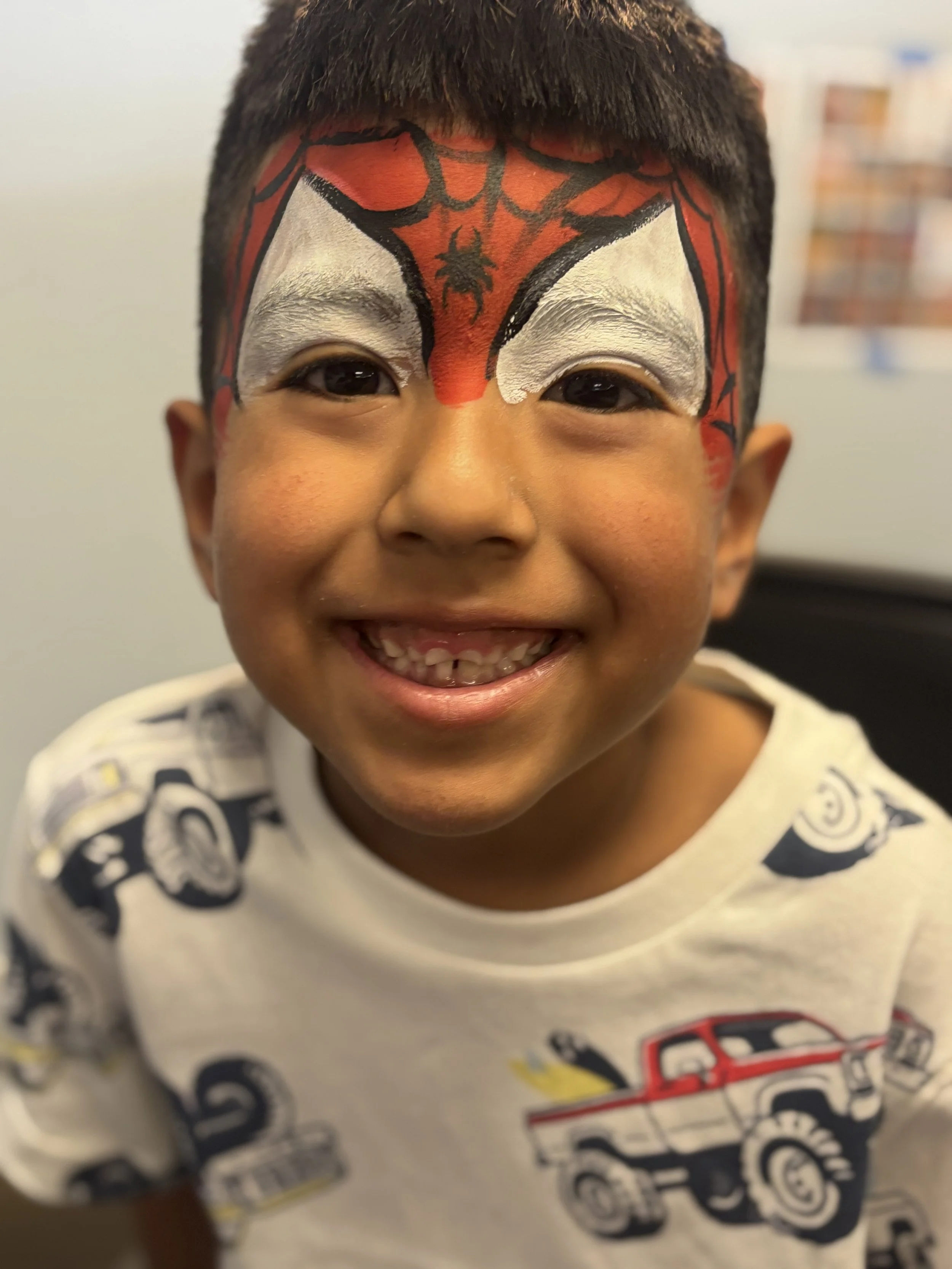 A young boy with face paint resembling Spider-Man, smiling and showing missing teeth, wearing a white shirt with robot and truck illustrations.