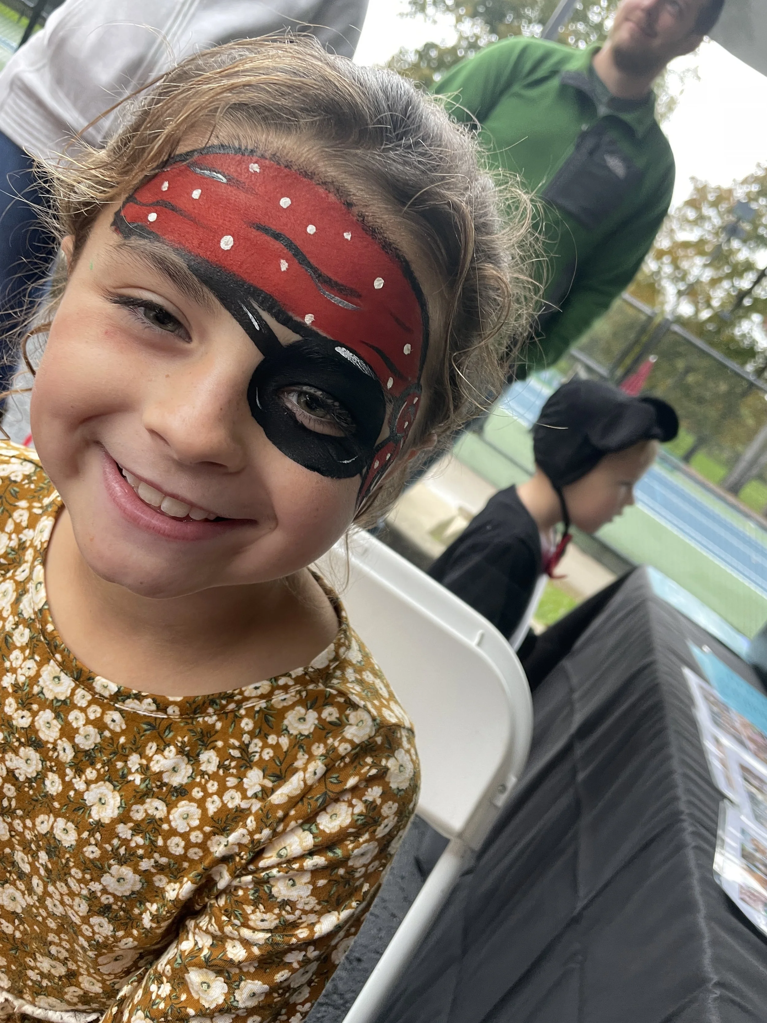 A young girl with face paint resembling a red and black butterfly or flower face mask, smiling at the camera. She is sitting outdoors near a tennis court, with a man in a green jacket and another girl in a helmet and black jacket visible in the backg