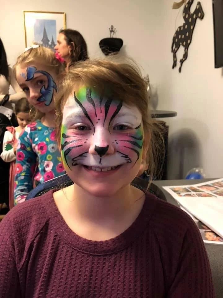 Child with face paint resembling a colorful tiger, smiling at the camera. In the background, other children and adults are visible, with one girl having face paint and wearing a floral shirt. The setting appears to be indoors with wall decor includin