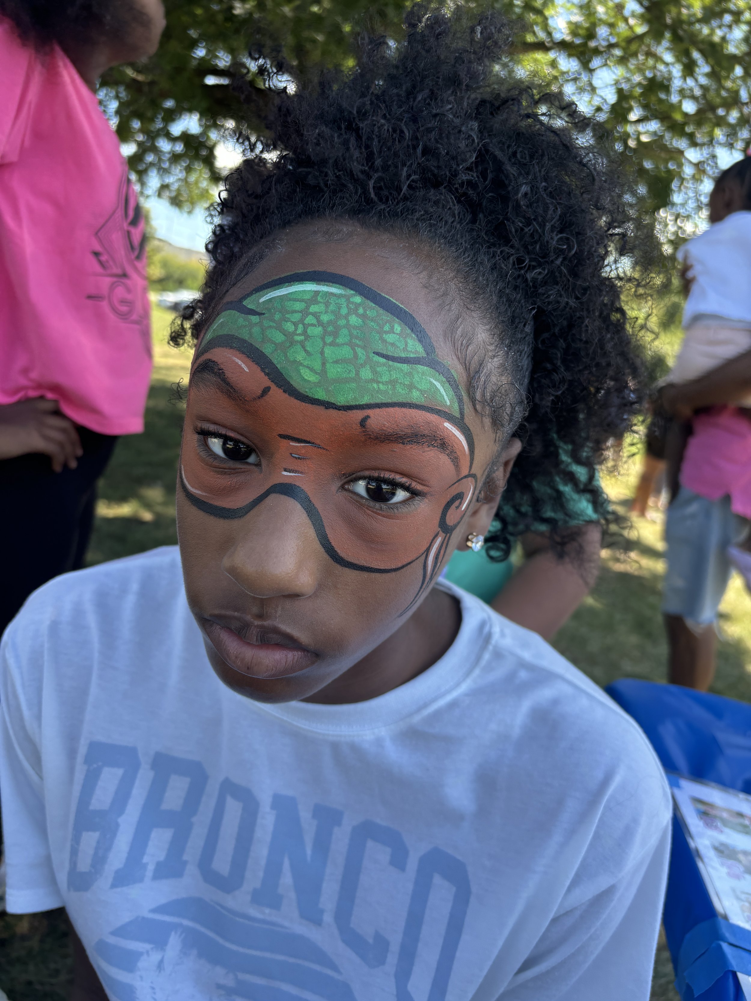 A young girl with curly hair and face paint resembling a turtle underwater scene, wearing a white t-shirt, outdoors in a park with other children around.