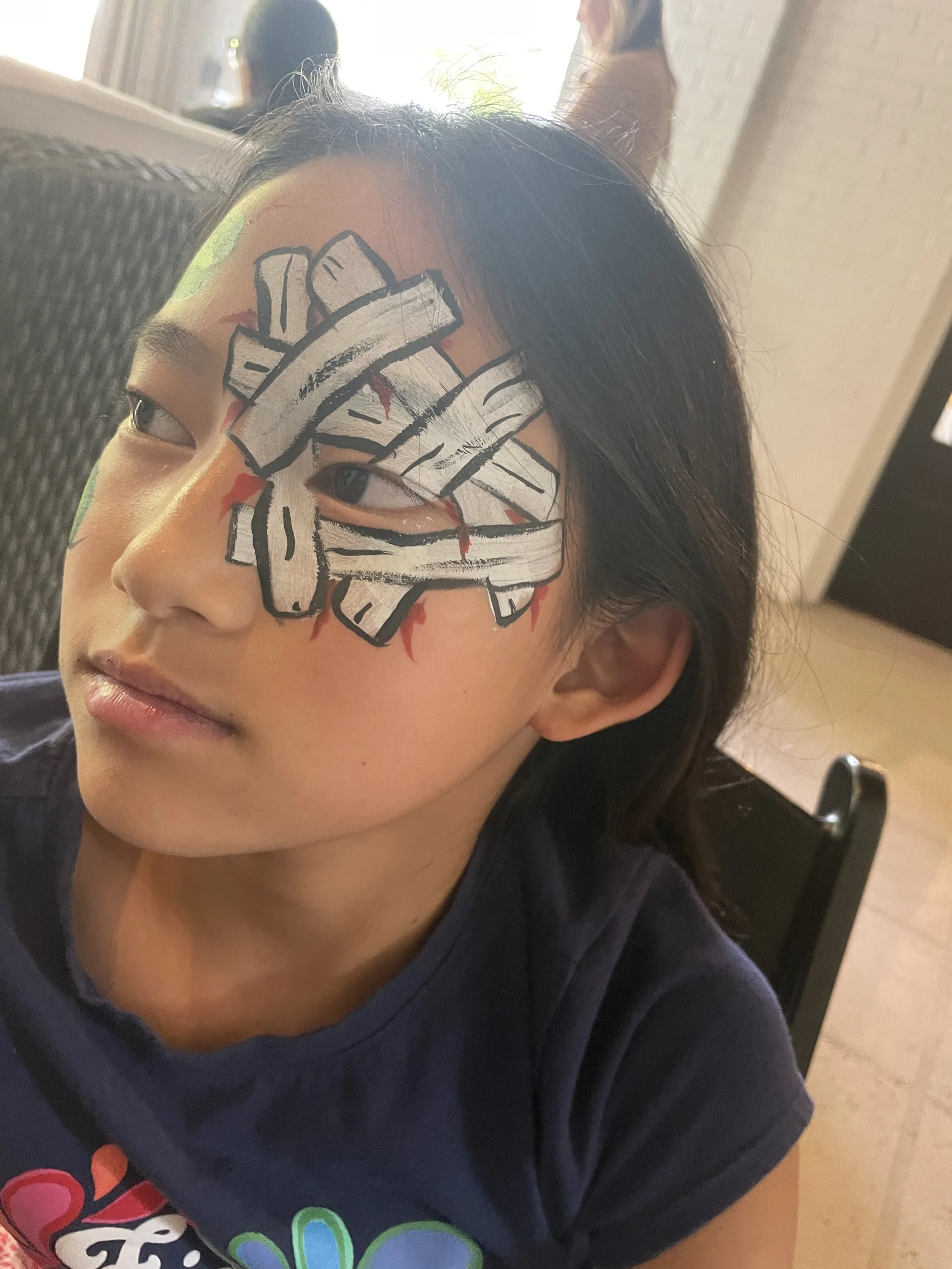 Young girl with a ninja mask painted around her eye, painted in black, white, and red, on her face. She has long dark hair and is sitting indoors, wearing a dark shirt.