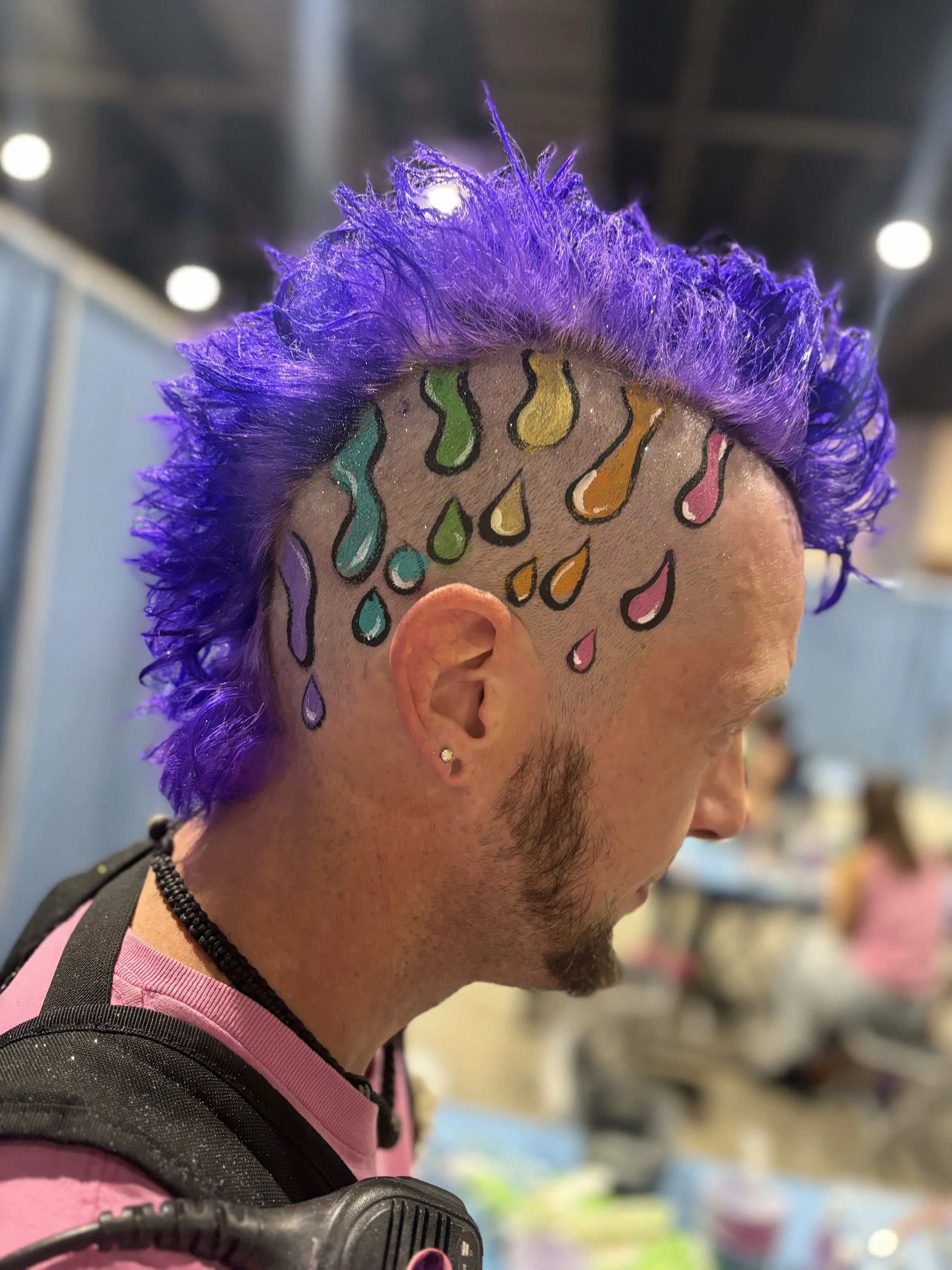 Person with purple spiked hair and colorful raindrop face paint, wearing a pink shirt and backpack.