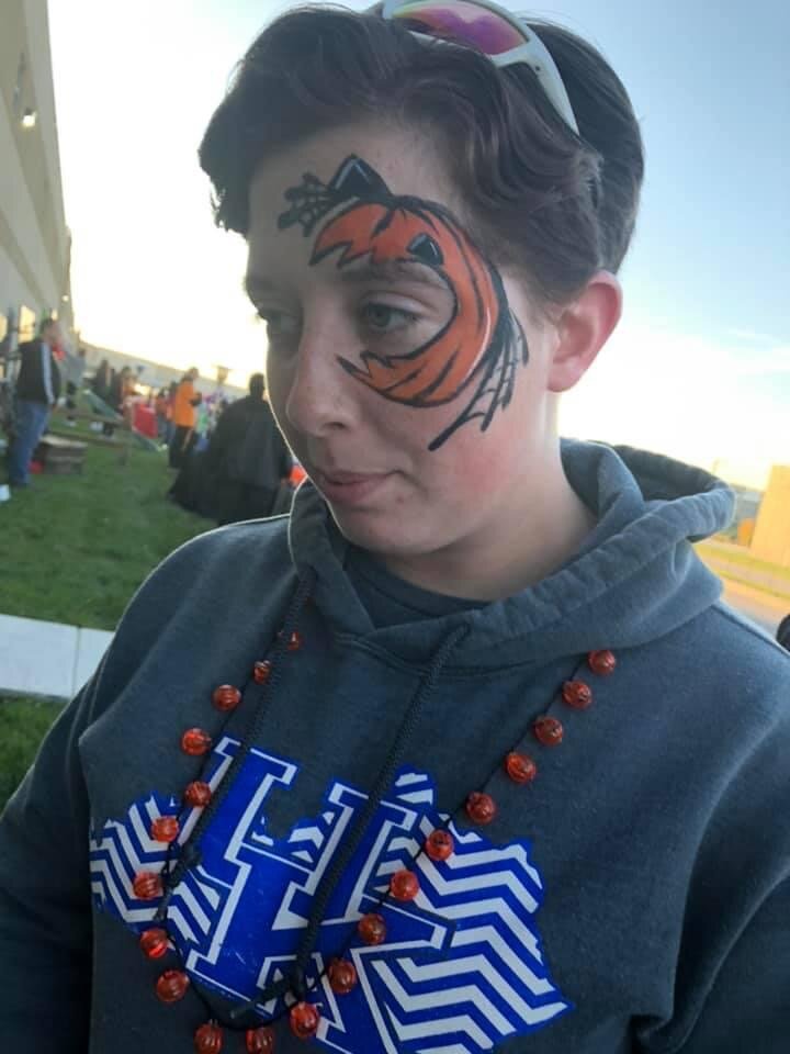 Teenager with face paint of a tiger on his right cheek, wearing sunglasses on his head, a gray hoodie, and an orange beaded necklace outdoors at daytime.