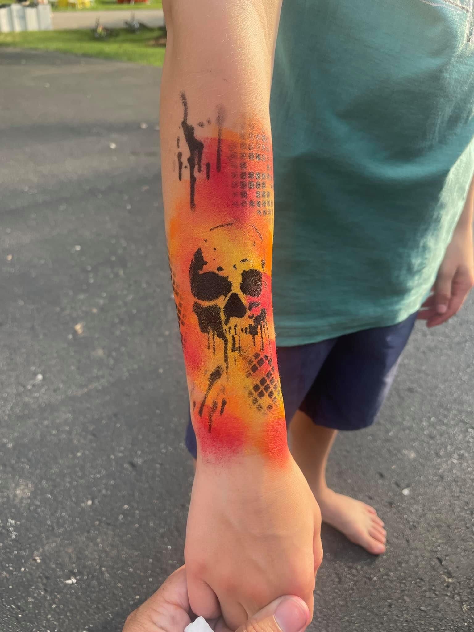 Tattoo of a black skull with drips and geometric patterns on a forearm, with a sunset background of orange and yellow hues.