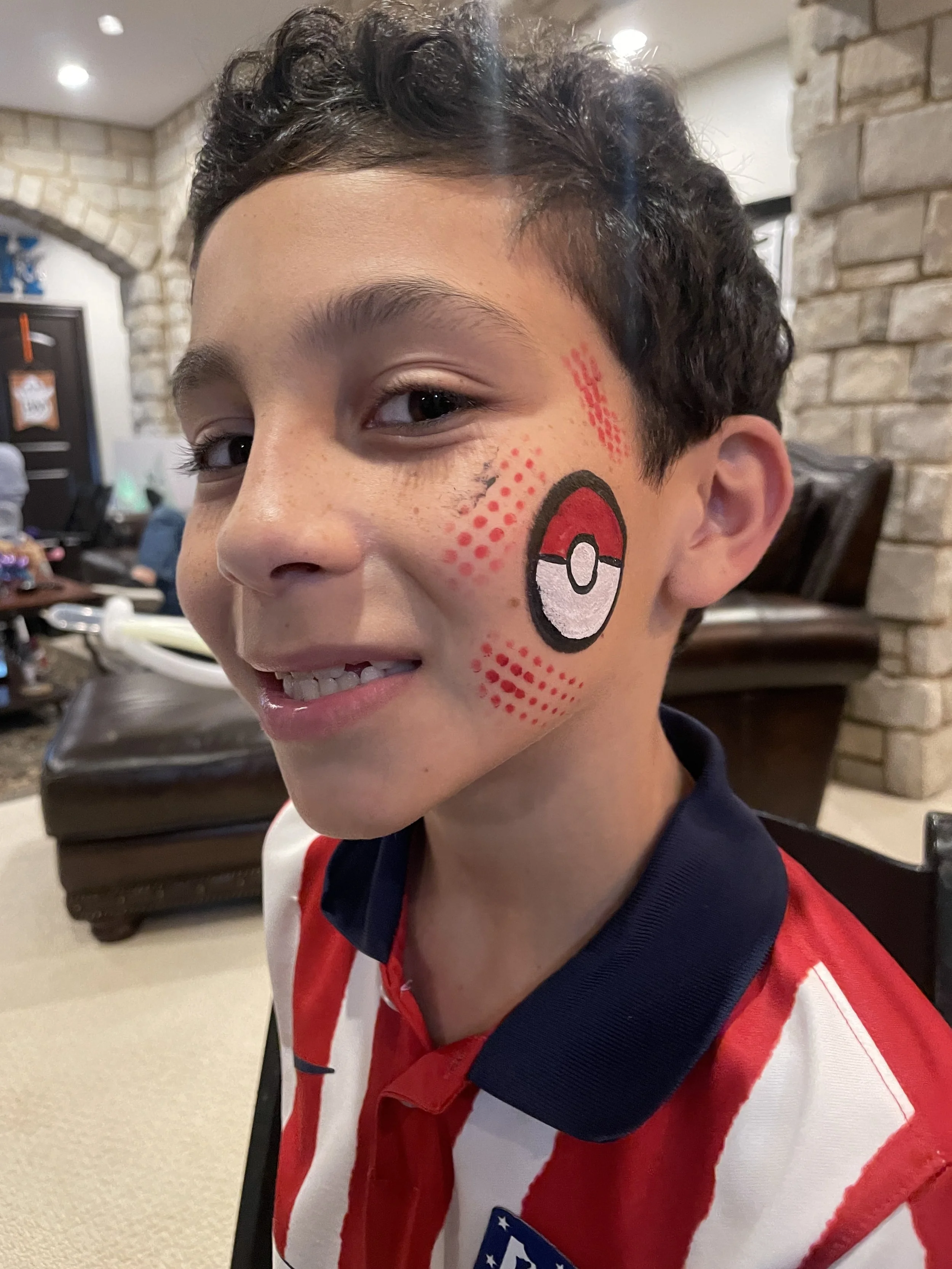 A young boy with a painted Poké Ball on his cheek, smiling, in a room with stone walls and people in the background.