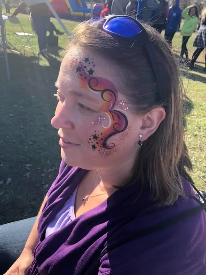A woman with face paint featuring swirls, stars, and sparkles, sitting outdoors at an event wearing sunglasses on her head and a purple jacket.