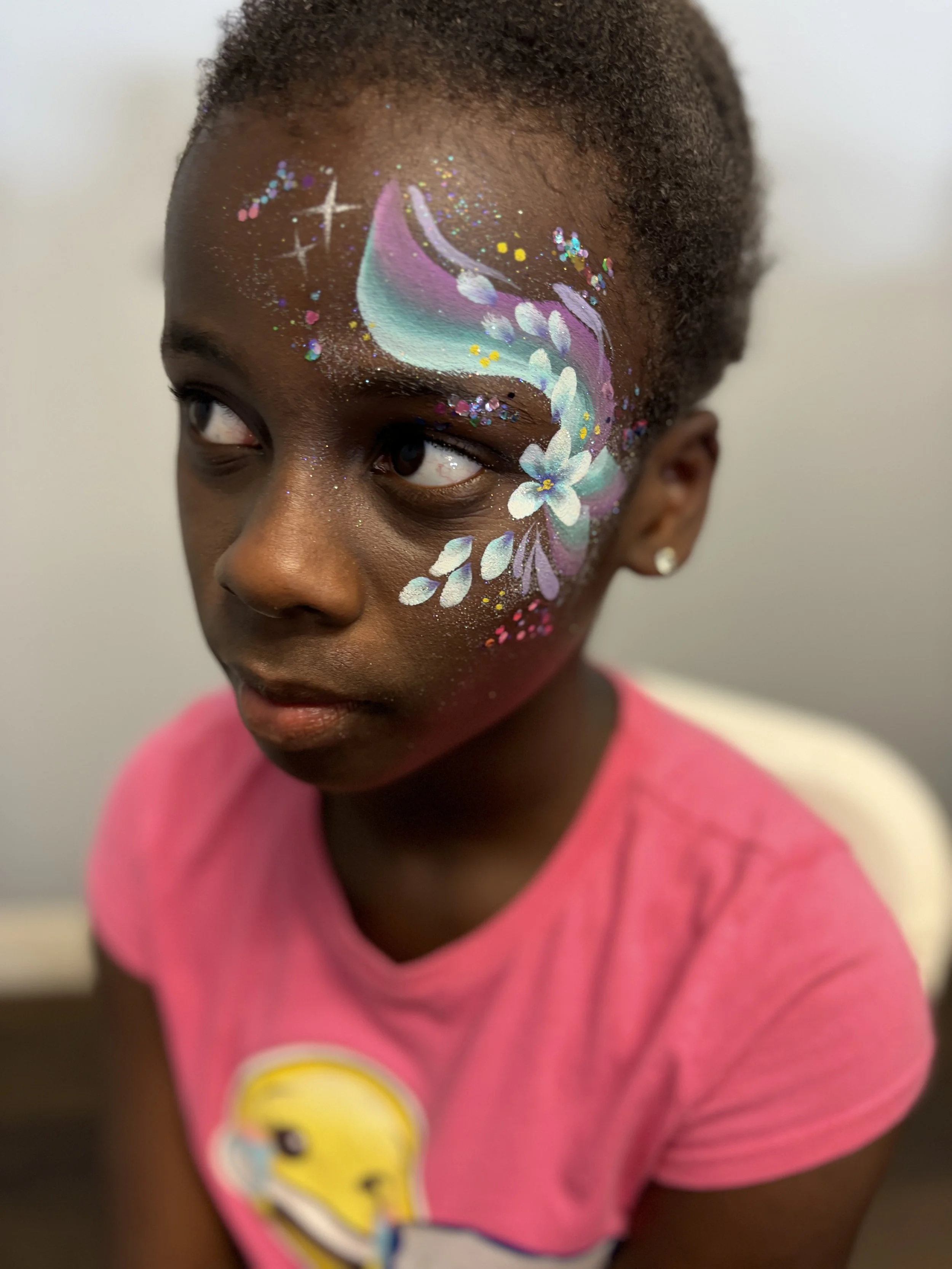Child with colorful space-themed face paint featuring a comet, stars, and flowers, wearing a pink shirt.