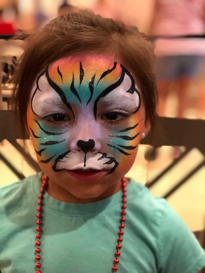Child with colorful rainbow face paint and black tiger stripe details, wearing a teal shirt and a red beaded necklace.